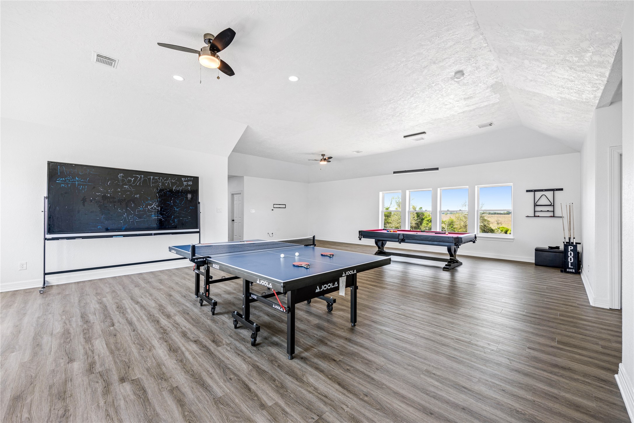 15015 House Road Cypress, TX 77433 - Photo 43 of 50 This spacious game room features sleek wood flooring, large windows with scenic views, a ceiling fan, and ample natural light. Equipped with a table tennis setup and a pool table, it's perfect for entertainment and leisure.