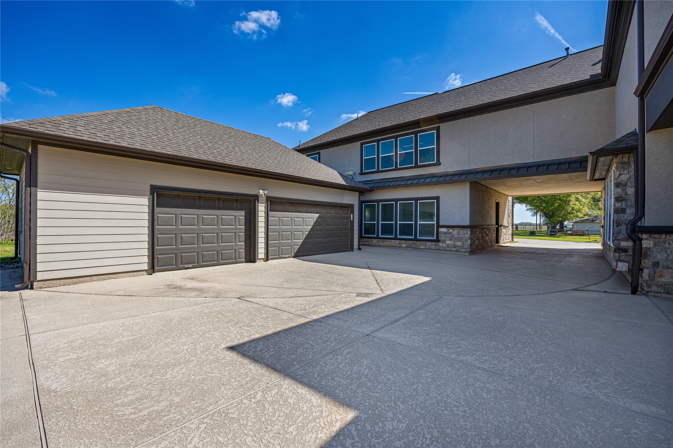 15015 House Road Cypress, TX 77433 - Photo 8 of 50 This photo showcases a spacious driveway leading to a three-car garage with modern doors. The exterior features a combination of stone and siding, with clean architectural lines and multiple large windows, allowing for ample natural light. The property includes an open area with a covered passageway, providing additional outdoor space and convenience.