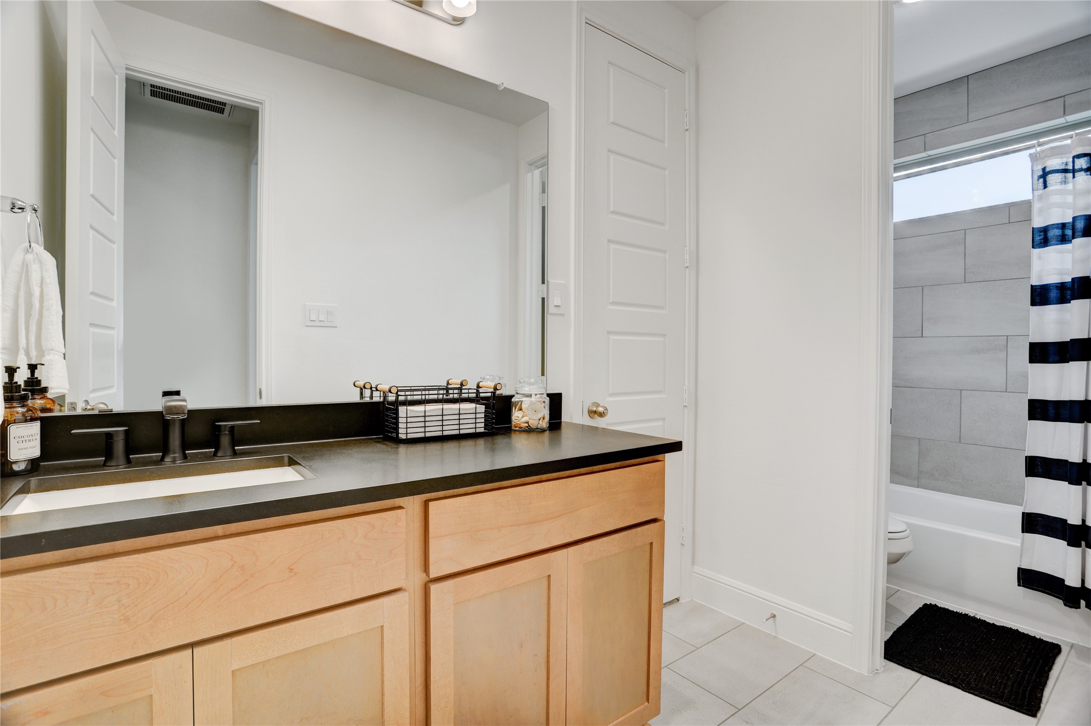 619 Rita Blanca Drive Webster, TX 77598 - Photo 23 of 44 This shared secondary bathroom features a modern design with a sleek black countertop and light wood cabinetry. It includes a large mirror above the sink, white walls, and a shower with gray tile offering a clean and functional layout.