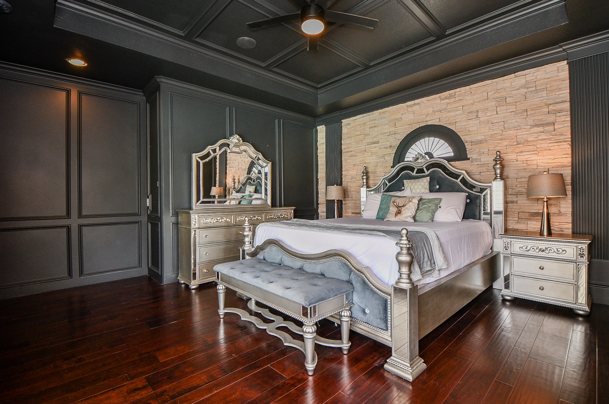 263 Wood Loop Street Houston, TX 77015 - Photo 29 of 45 Elegant bedroom featuring a luxurious bed with ornate detailing, a matching dresser, and nightstands. The room has rich dark wood flooring, a stylish coffered ceiling, and a stone accent wall. Warm lighting and a soft bench enhance the sophisticated ambiance.