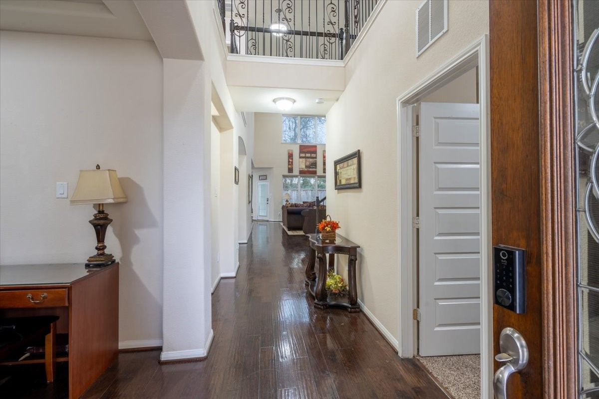 This photo showcases a welcoming entryway with dark hardwood floors, a high ceiling, and a view into a cozy living area. There's a decorative lamp on a wooden table to the left and an open door leading to a room on the right. A staircase with wrought iron railing is visible above, adding elegance to the space.