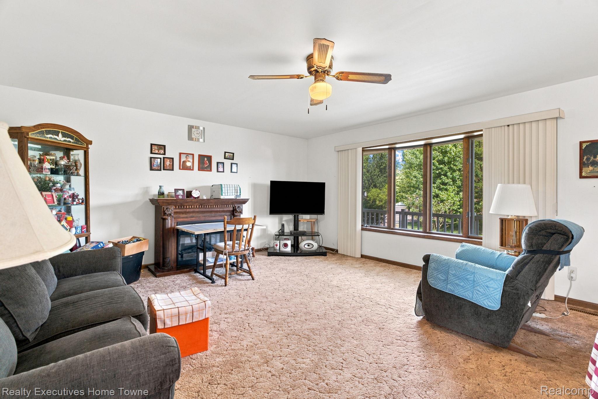 959 Braidwood Road Memphis, MI 48041 - Photo 16 of 58 Spacious living area featuring a decorative wood-mantle fireplace, a large picture window with vertical blinds, a ceiling fan with integrated lighting, and wall-to-wall carpeting