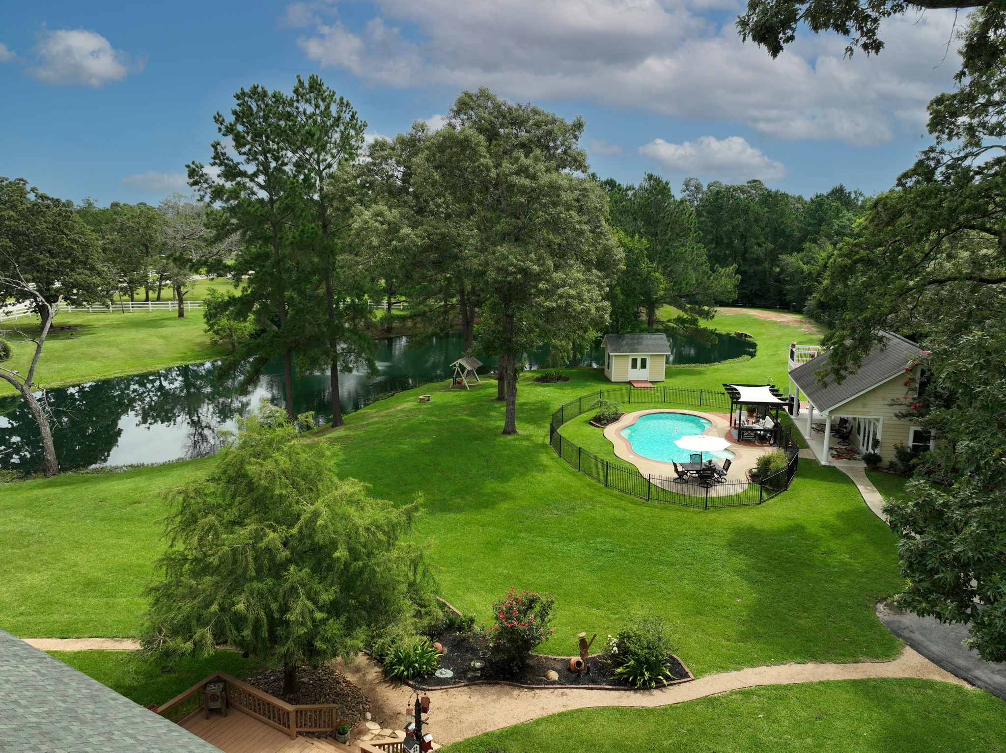 34310 High Point Drive Magnolia, TX 77355 - Photo 25 of 48 This aerial view shows lush greenery, a serene pond, and a fenced-in pool area. The charming guest house with a pergola offers a perfect spot for relaxation, surrounded by mature trees and manicured landscaping. Ideal for those seeking a peaceful retreat.