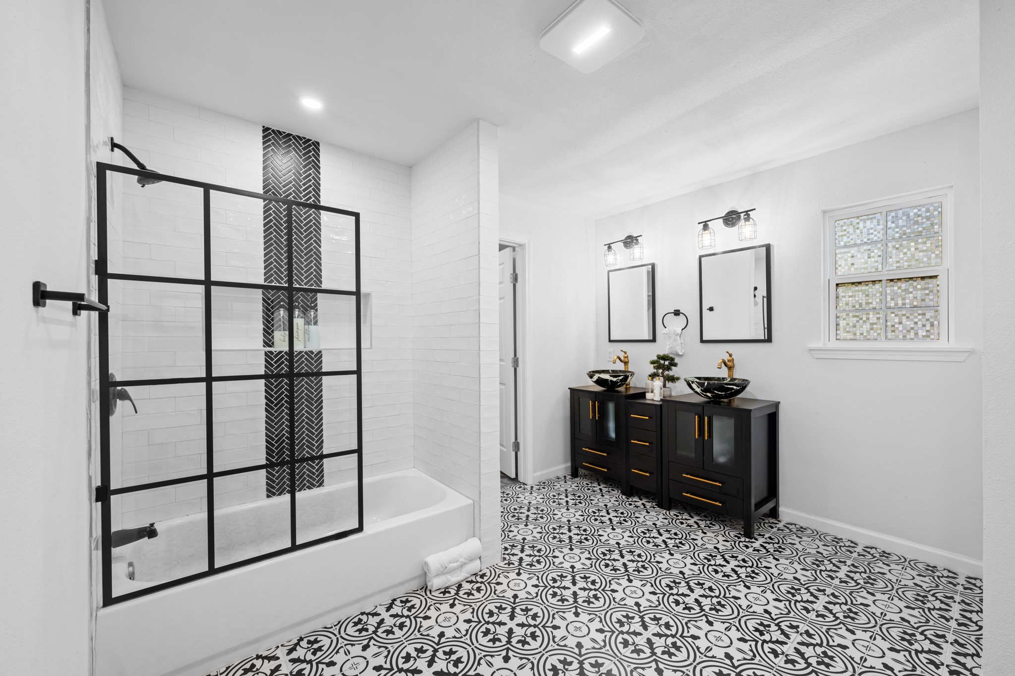 729 Kyle Angleton, TX 77515 - Photo 22 of 36 This bathroom features a modern design with a black-and-white color scheme. It includes a sleek shower with a glass partition, a double vanity with vessel sinks, and stylish patterned floor tiles. Ample natural light enters through a window, enhancing the bright and airy feel.
