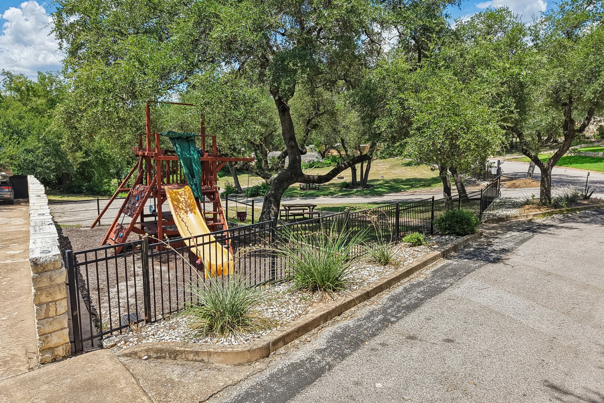 13101 Travis View Loop Austin, TX 78732 - Photo 21 of 27 A private playground area with a slide and swing set, framed by lush trees and a stone wall.