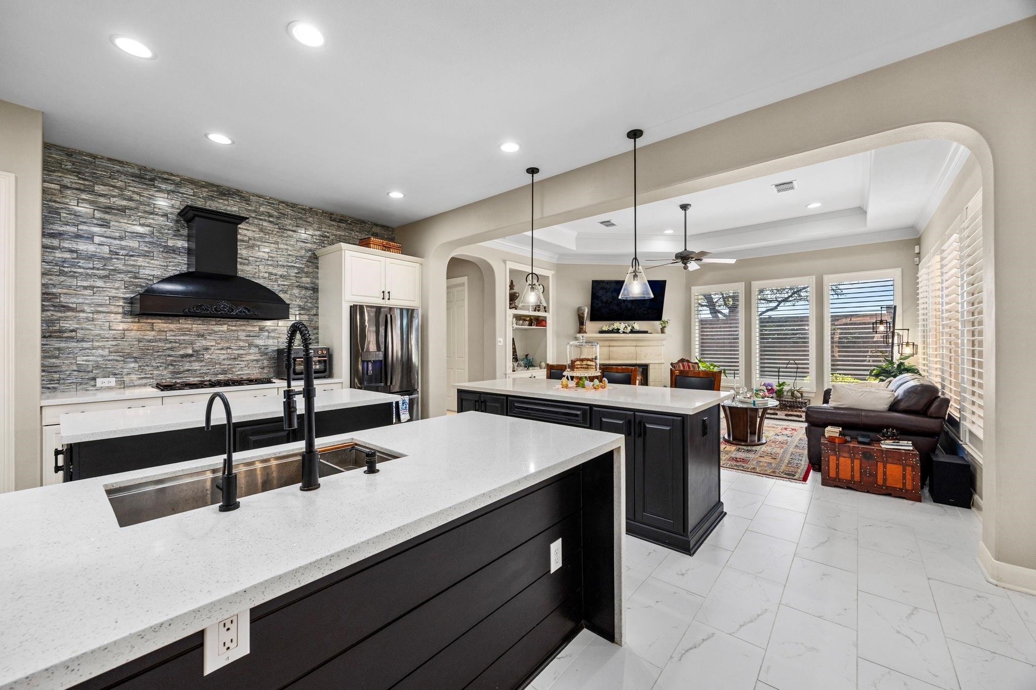 11413 Gallant Ridge Lane Houston, TX 77082 - Photo 20 of 50 Photo showcases a modern, open-concept kitchen and living area. The kitchen features sleek white countertops, dark cabinetry, a stylish backsplash, and new appliances. It opens to a cozy living room with large windows, allowing natural light to fill the space. The room is accented with a fireplace and elegant lighting fixtures, creating a warm and inviting atmosphere.