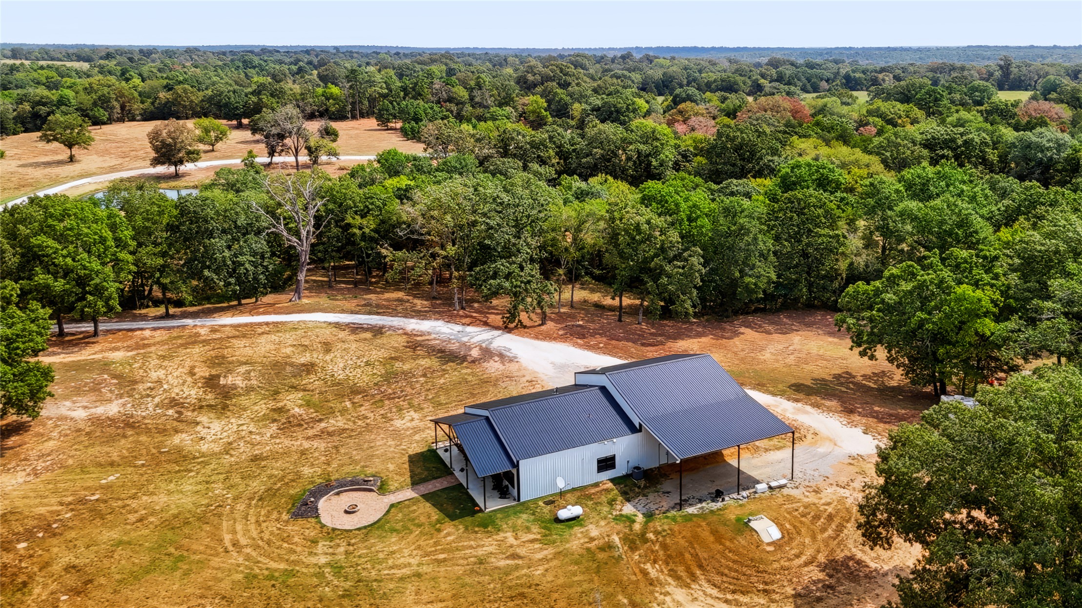 3607 South Half Bank Road Atoka, OK 74525 - Photo 58 of 82 Single-story residence with a dark metal roof, covered carport, fire pit area, and extensive surrounding treeline