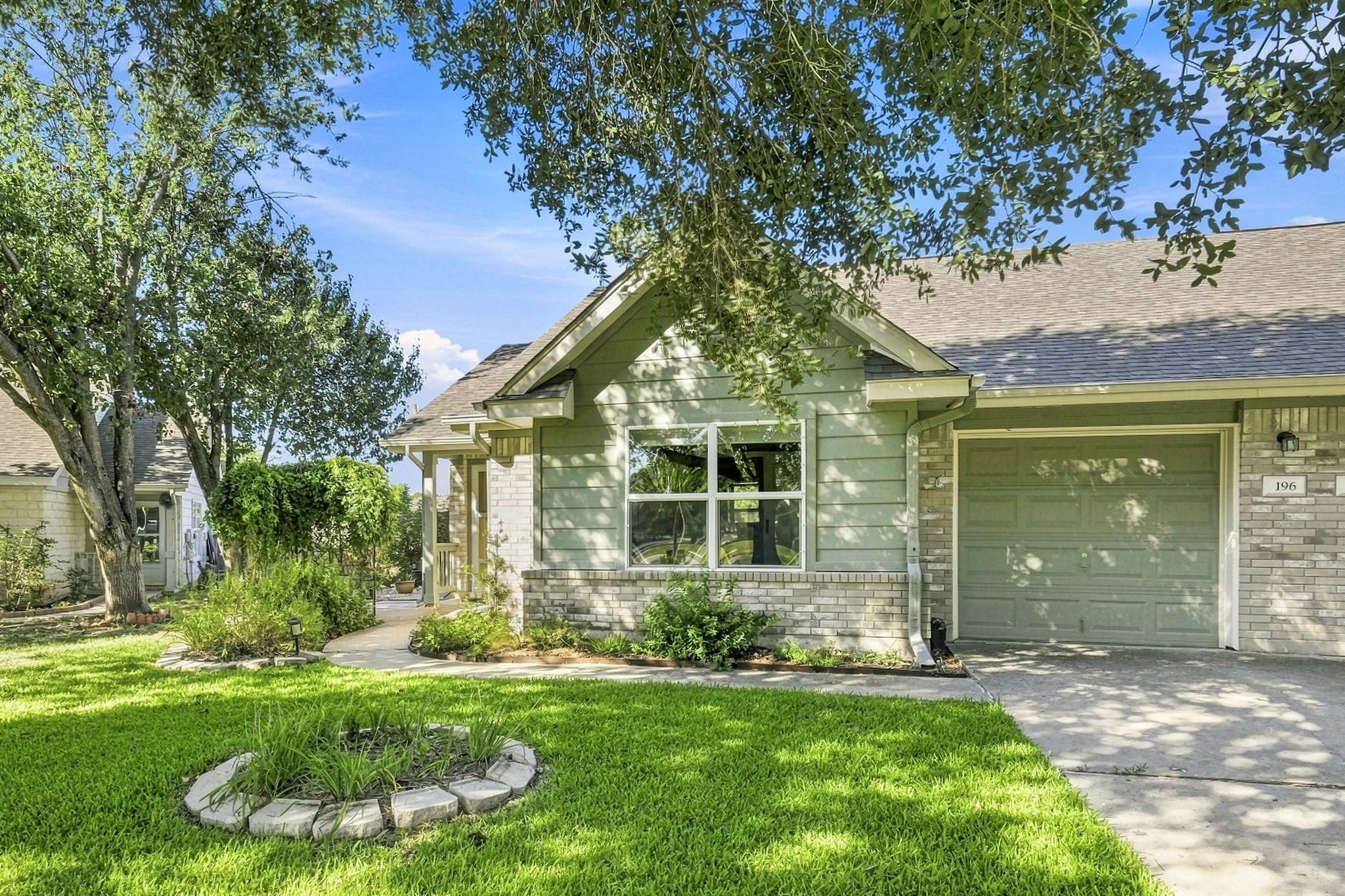 Mature trees shade a manicured front lawn leading to a brick-accented exterior and garage.