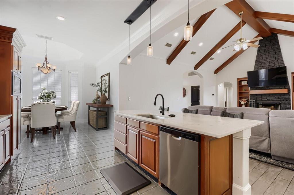 603 Ash Street Pilot Point, TX 76258 - Photo 12 of 28 Spacious open-concept interior featuring vaulted ceilings with exposed wood beams, a central kitchen island with a built-in stainless steel dishwasher, a fireplace with a stacked stone facade, and wood-finish cabinetry