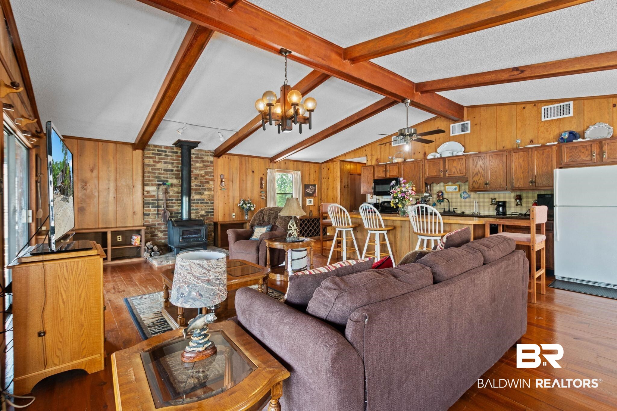 8153 Bryant's Lndg Road Stockton, AL 36579 - Photo 20 of 87 Vaulted ceiling with exposed wood beams, wood-finish flooring, wood-paneled walls, brick accent wall with a wood-burning stove, and a kitchen island with seating