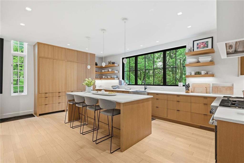 810 Persimmon Road Sewickley, PA 15143 - Photo 7 of 49 The chef's kitchen features flat-panel quarter-sawn white oak cabinetry, thick marble countertops, a generous center island with seating, and open wood shelving framed by a dramatic oversized window.