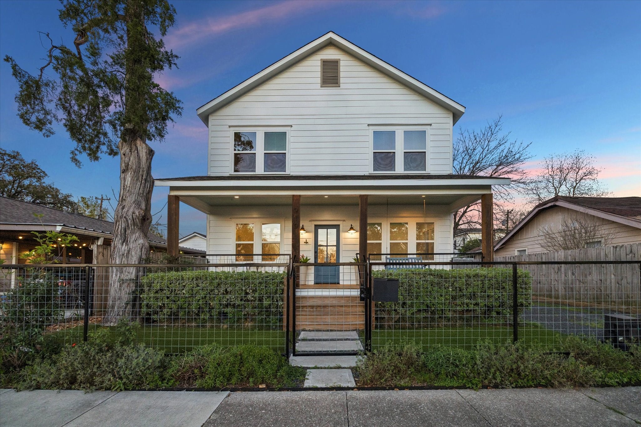Charming two-story home with a welcoming front porch, surrounded by greenery and enclosed with a modern fence.