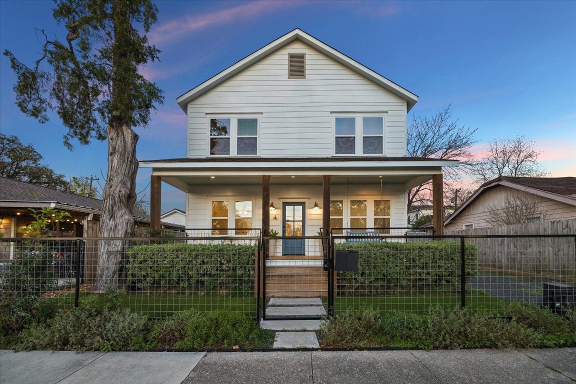 Charming two-story home with a welcoming front porch, surrounded by greenery and enclosed with a modern fence.