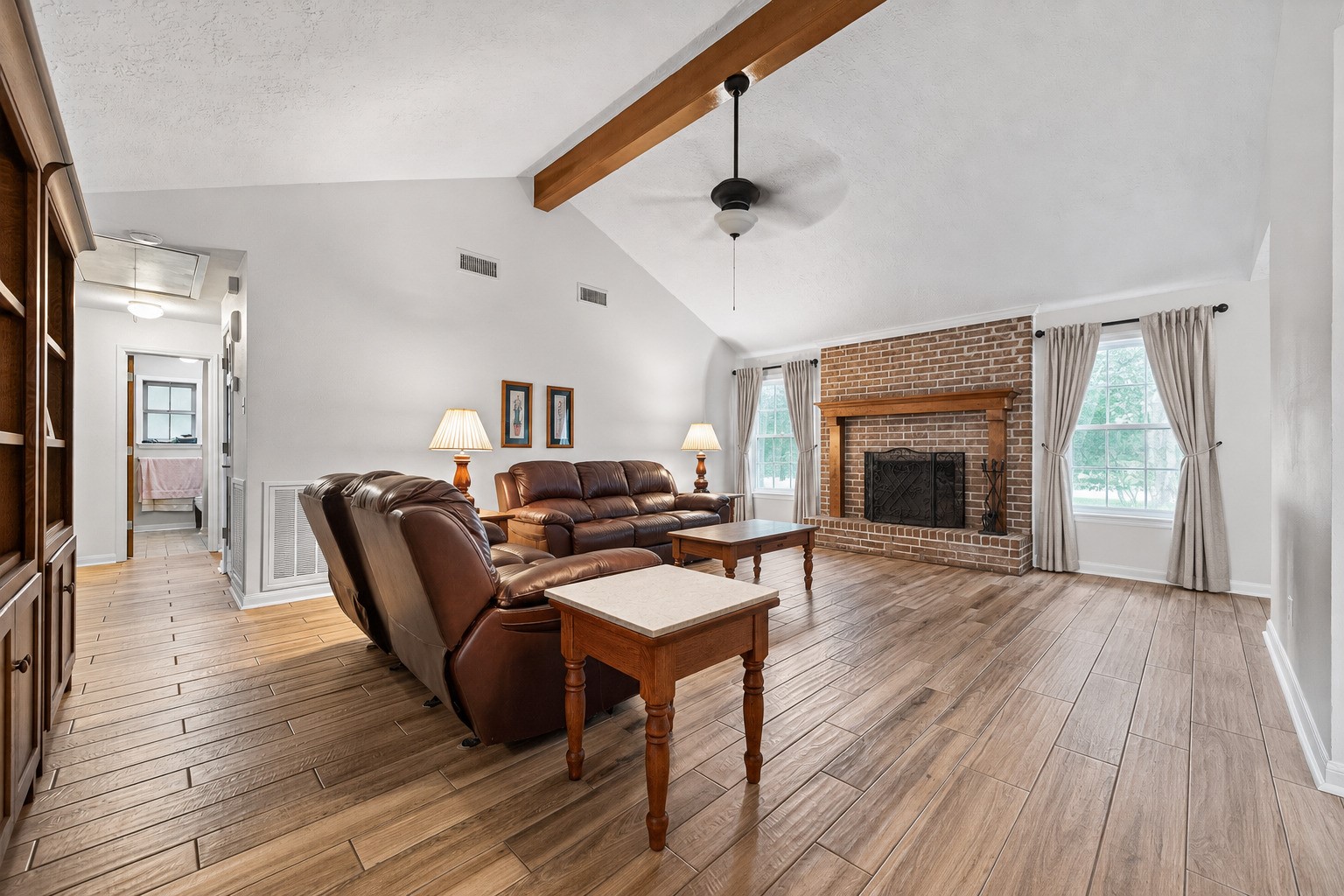 1004 Wagon Road Simonton, TX 77485 - Photo 5 of 40 This spacious living room impresses with soaring vaulted ceilings accented by a rustic wood beam and a ceiling fan that keeps the space comfortable year-round. At the heart of the room stands a classic full brick fireplace with a warm wood mantel, serving as a stunning focal point. Wood-look tile flooring flows throughout, adding warmth and continuity to this generous and inviting space.