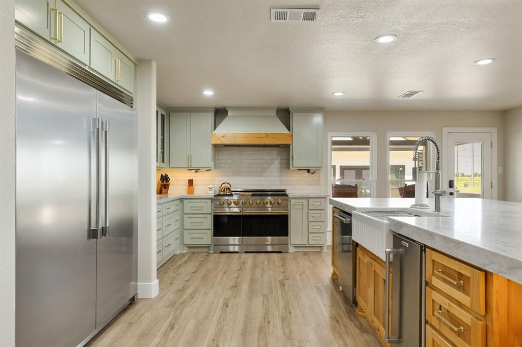 12240 Wellington Road Ponder, TX 76259 - Photo 11 of 40 Gourmet kitchen featuring a large stainless steel refrigerator, professional-grade range, custom light green cabinetry, wood-finish flooring, and a spacious island with a farmhouse sink