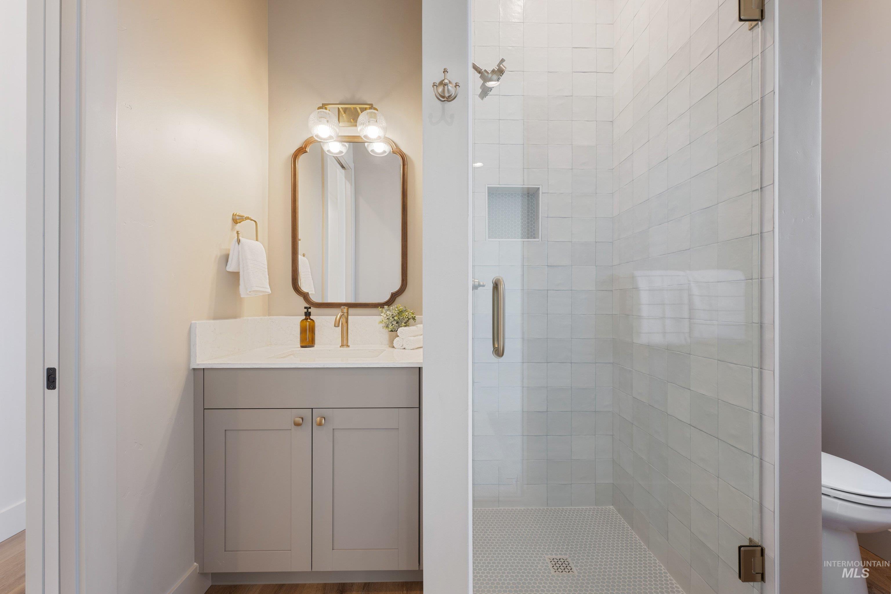 24821 Ardmore Court Caldwell, ID 83607 - Photo 28 of 43 Bathroom featuring a glass-enclosed shower with white tiled walls and hexagonal mosaic floor, a vanity with a light gray cabinet, white countertop, and gold-finish hardware, and a decorative mirror with an overhead light fixture