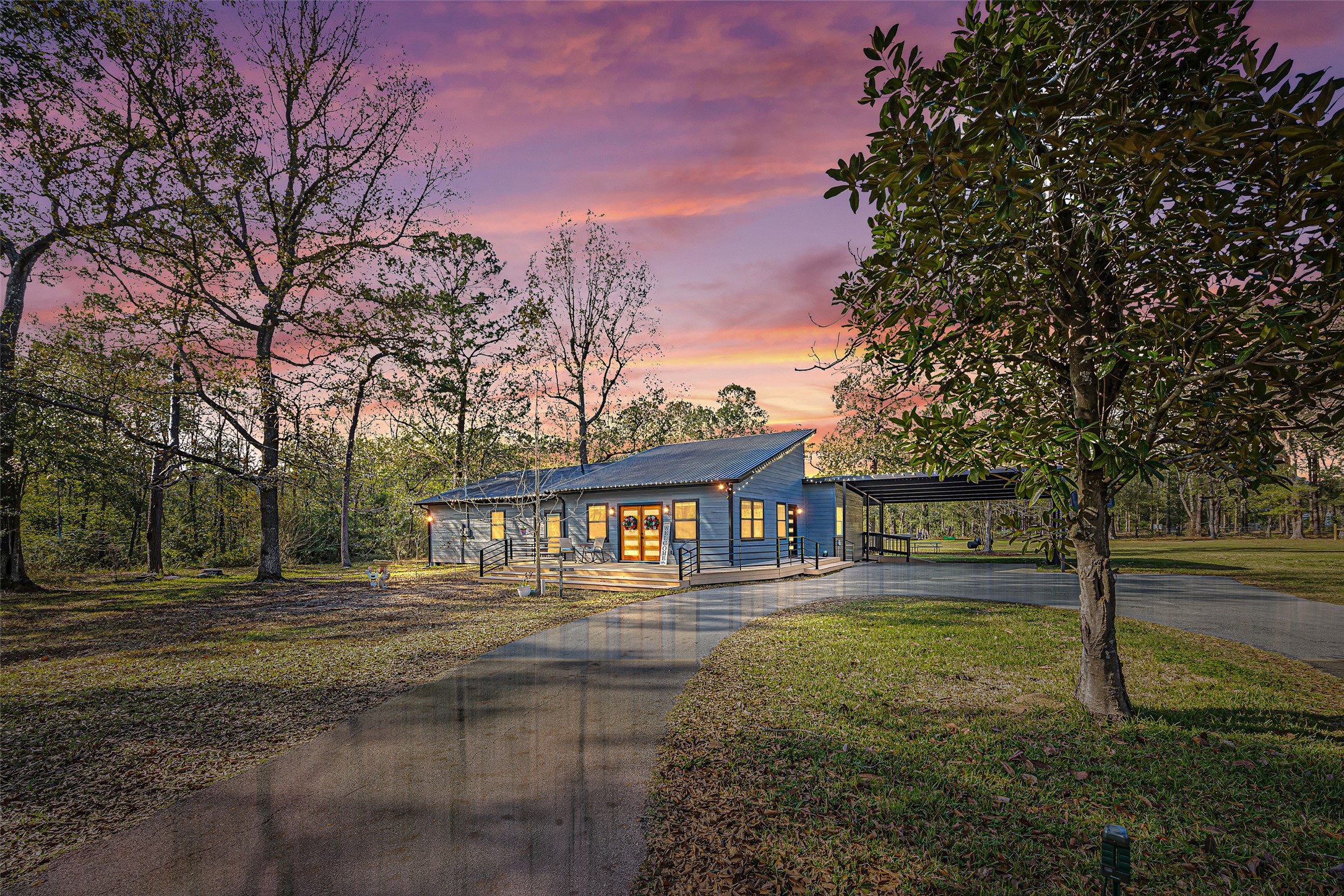 15080 Usher Lane Splendora, TX 77372 - Photo 1 of 44 This photo showcases a charming single-story home set in a wooded area, featuring a spacious driveway and a carport. The house has a modern design with large windows and a welcoming porch, surrounded by lush greenery, under a picturesque sky.