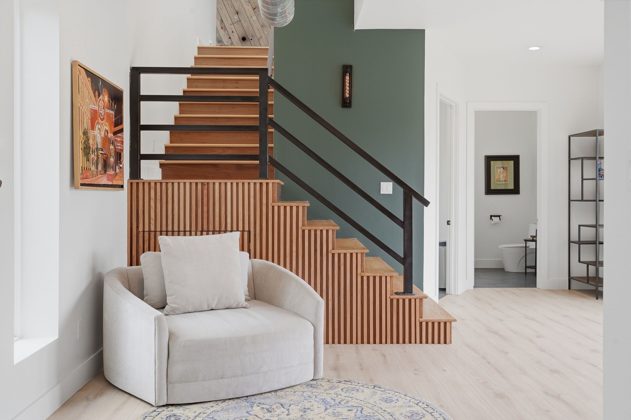 2743 Bobville Road Montgomery, TX 77316 - Photo 27 of 50 This photo showcases a modern interior with a stylish wooden staircase (with built in storage) featuring black railings. The neutral-toned room includes a cozy armchair and decorative elements like a wall painting and a minimalist shelving unit. The space feels open and inviting, with light wood flooring and a small glimpse into an adjacent room.