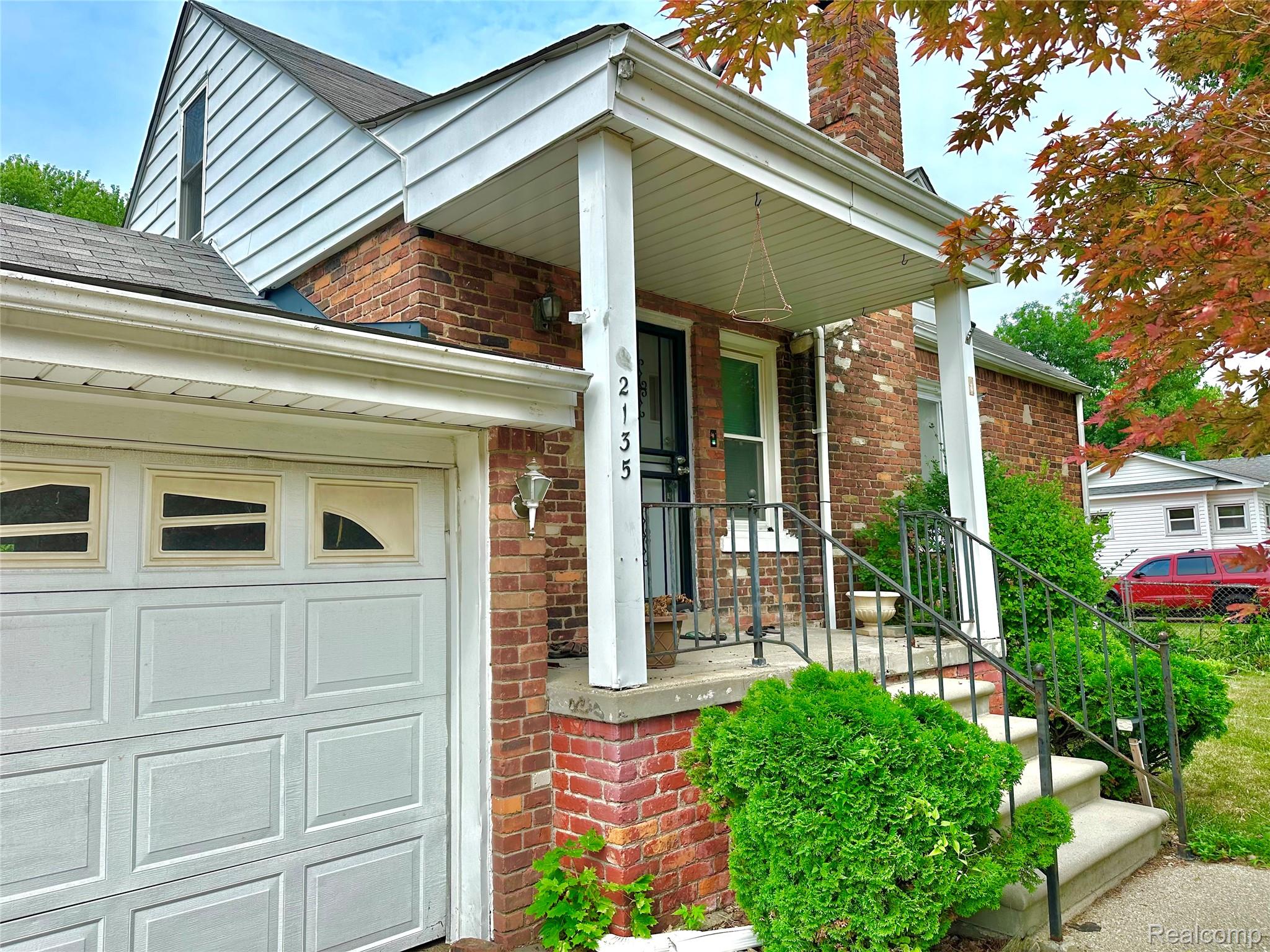 Brick exterior featuring a covered entry, white garage door, concrete steps with metal railings, and established landscaping