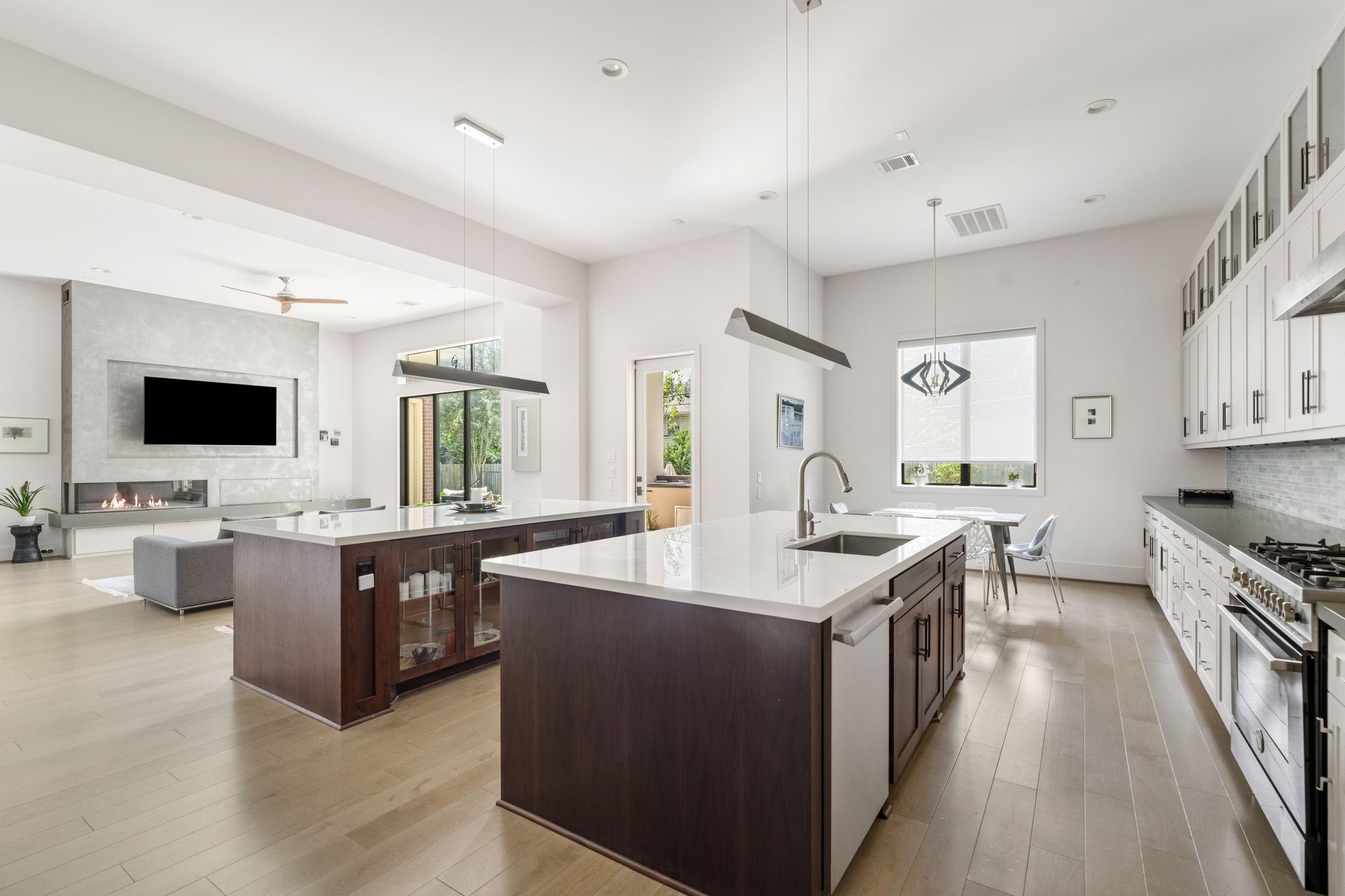 5427 Maple Street Houston, TX 77096 - Photo 18 of 46 The kitchen spans an impressive width, anchored by two substantial islands with dark wood bases and light stone countertops. The open sightline extends into the living area, where the fireplace wall is visible in the distance.