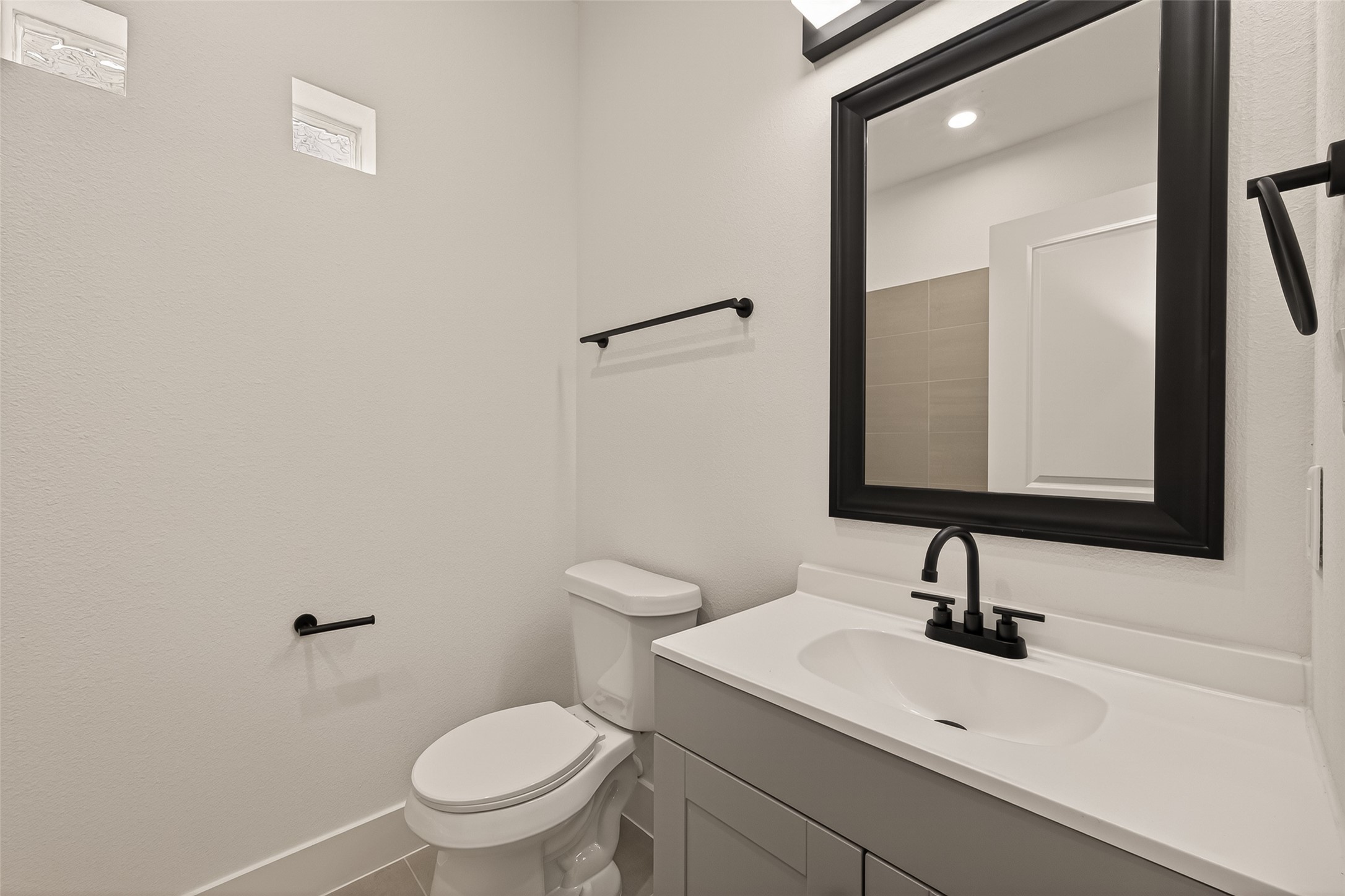 6808 Madrid Street Houston, TX 77021 - Photo 20 of 31 This bathroom features a clean, modern design with a white vanity and black fixtures. It includes a toilet and a large mirror above the sink, complemented by neutral tile flooring and two small windows for natural light.