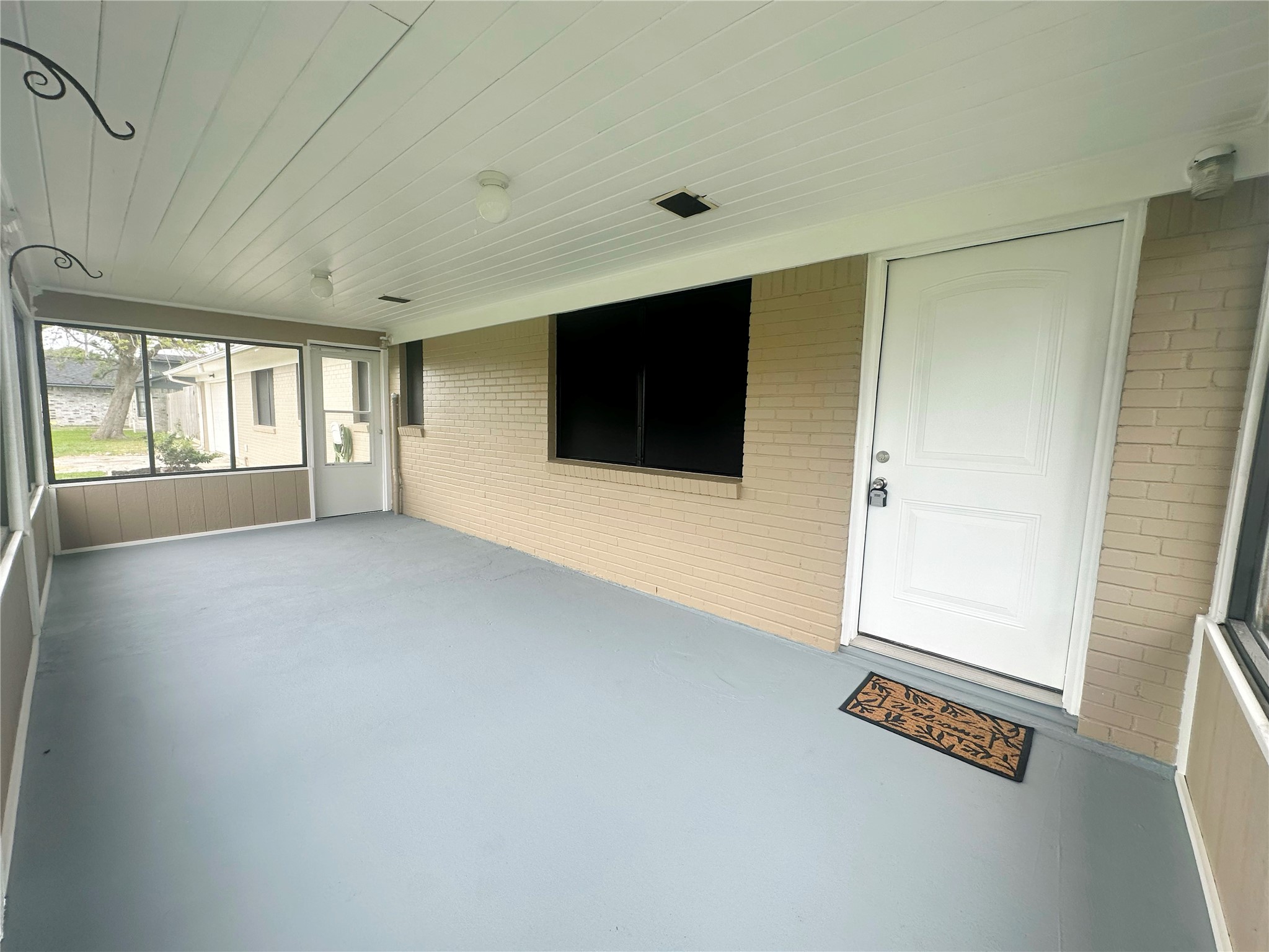 130 Bay Ridge Drive Baytown, TX 77523 - Photo 3 of 20 This image shows a spacious enclosed porch with a light ceiling, beige brick walls, and a clean, gray floor. Large windows provide ample natural light, and there’s a white entrance door with a welcome mat.
