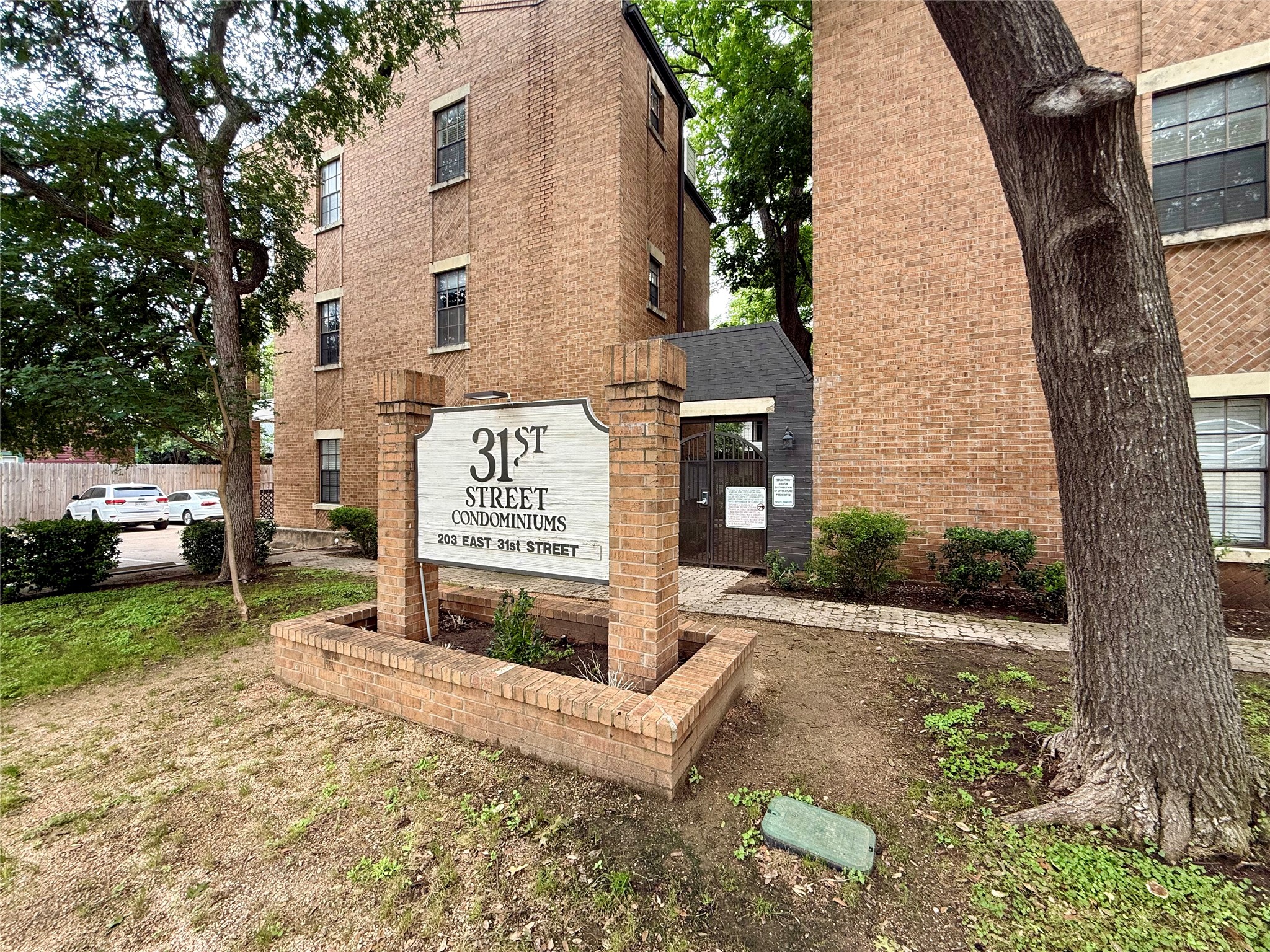 203 East 31st Street, Unit 101 Austin, TX 78705 - Photo 3 of 24 Brick condominium building with a prominent entryway sign, bordered by established trees and a paved walkway