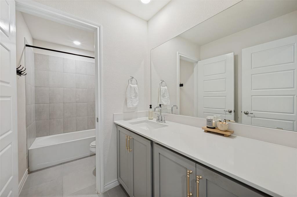 9828 Quail Pointe Road Aubrey, TX 76227 - Photo 15 of 23 Full-width vanity mirror, gray shaker-style cabinetry with gold-tone hardware, white countertop, undermount sink, and tiled tub surround