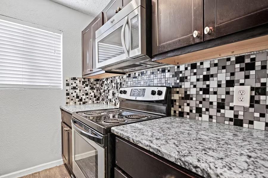 Kitchen featuring a textured wall, tasteful backsplash, appliances with stainless steel finishes, dark brown cabinetry, and light stone countertops
