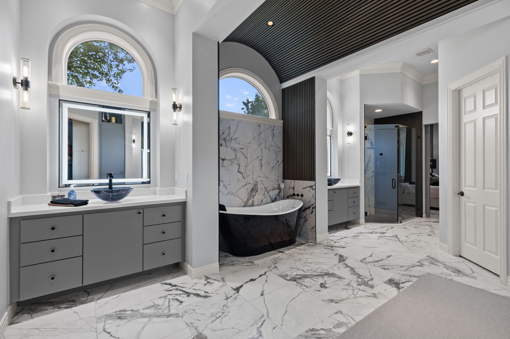 39 Maymont Way Spring, TX 77382 - Photo 22 of 50 This luxurious bathroom features elegant marble flooring, a freestanding black bathtub, modern dual vanities with vessel sinks, and large arched windows for ample natural light. The sleek, minimalistic design is complemented by contemporary lighting and a spacious glass shower enclosure.