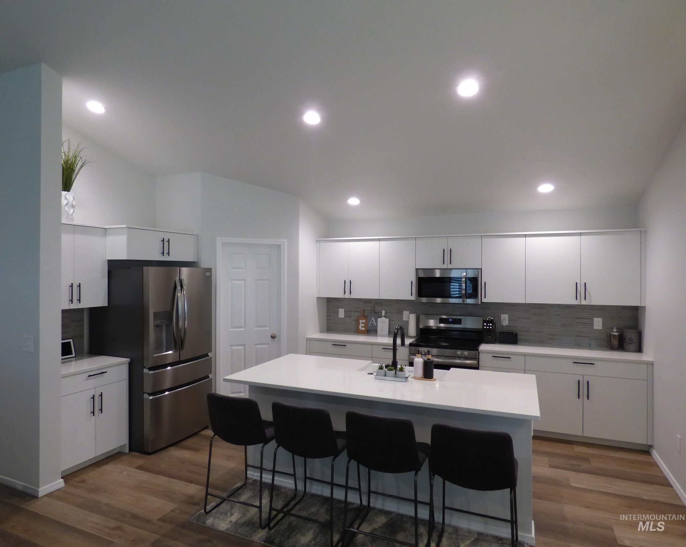 2708 East Buckaroo Drive Kuna, ID 83634 - Photo 5 of 48 Modern kitchen featuring a central island with a solid surface countertop, white cabinetry with dark hardware, stainless steel appliances, a tile backsplash, and wood-finish flooring