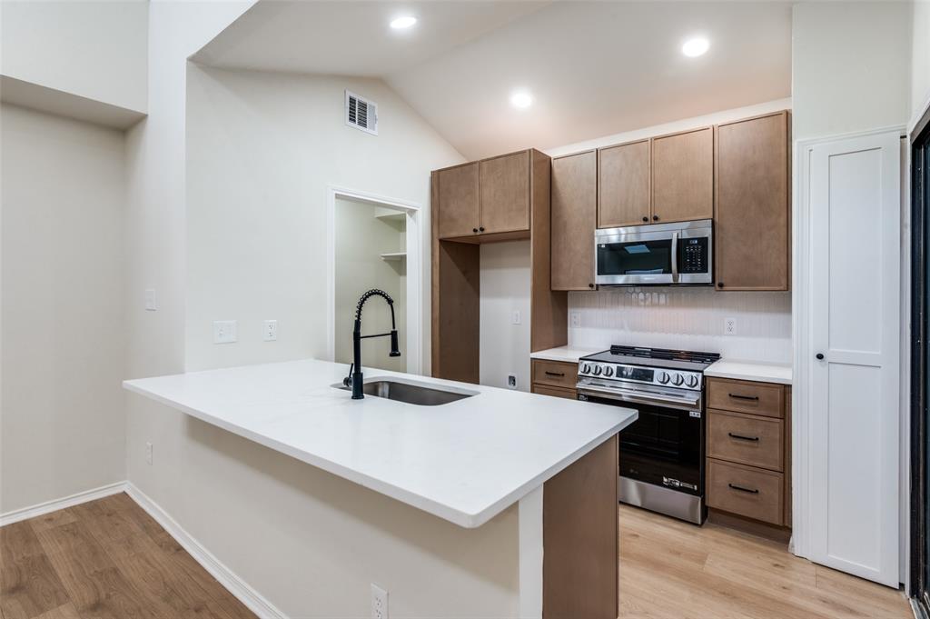 17490 Meandering Way, Unit 2202 Dallas, TX 75252 - Photo 14 of 24 Kitchen featuring an island with a built-in sink and matte black faucet, wood-finish cabinetry, stainless steel oven and microwave, white subway tile backsplash, and wood-finish flooring
