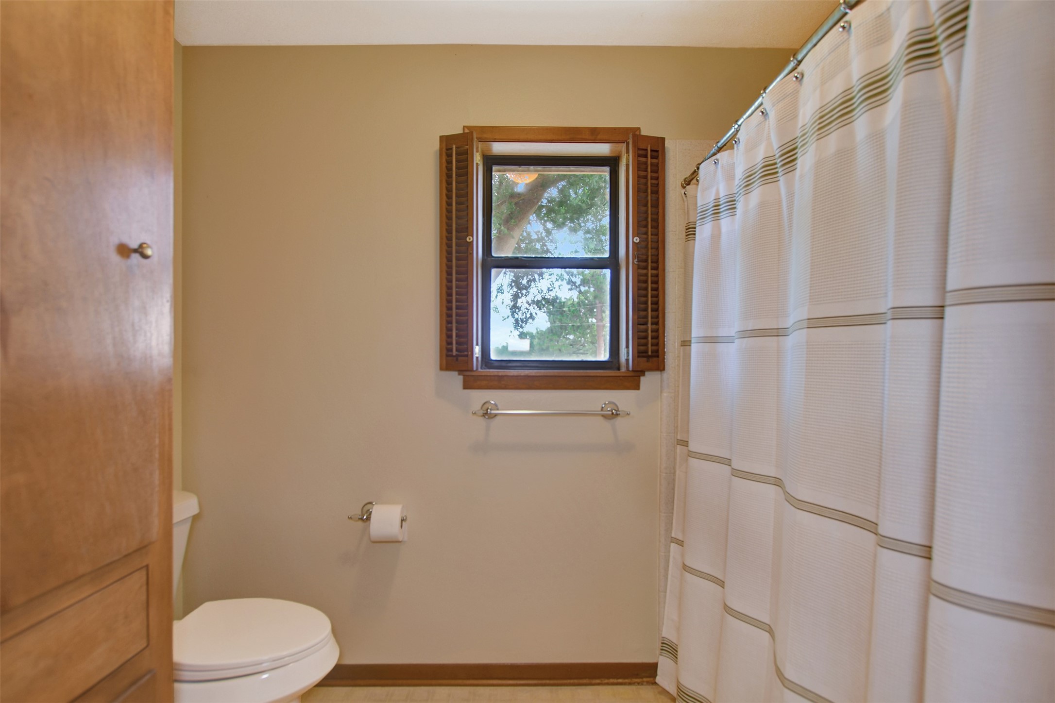341 South S Loop Bellville, TX 77418 - Photo 24 of 33 Cozy bathroom with neutral tones, featuring a window with wooden shutters, a shower with a striped curtain, and a toilet. A cabinet provides additional storage.