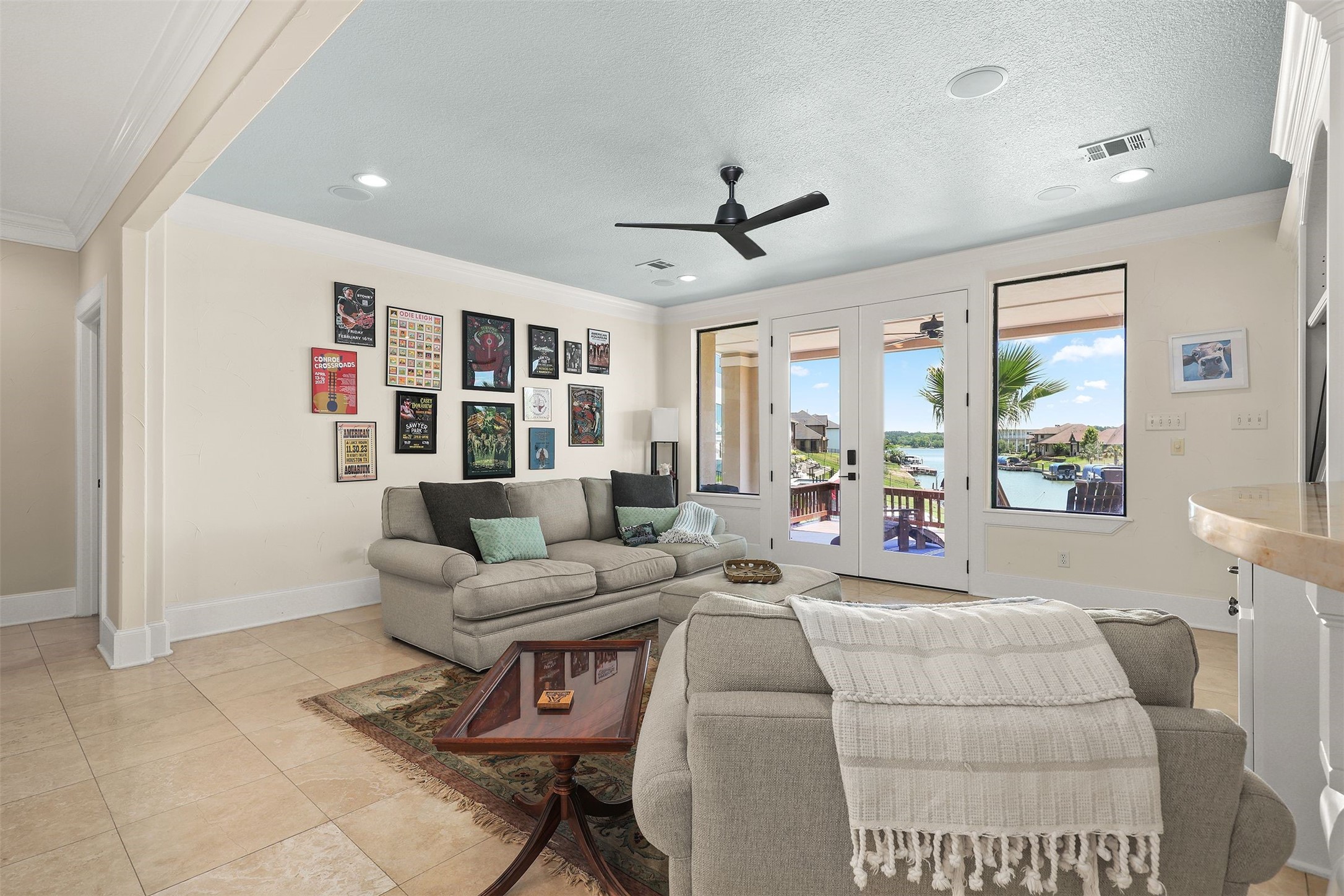 10896 Decatur Street Willis, TX 77318 - Photo 38 of 50 This bright living room, with its large windows and glass doors showcasing a stunning waterfront view, offers a cozy and inviting atmosphere with comfortable seating, tile flooring, a ceiling fan, and tasteful wall art.