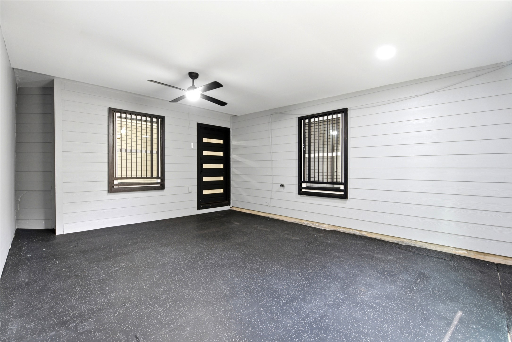 9403 Spaulding Street Houston, TX 77016 - Photo 31 of 50 This space features a modern, enclosed area with light gray siding and sleek black-framed windows. It includes a stylish black door with horizontal glass panels and a ceiling fan, providing a clean and versatile setting.