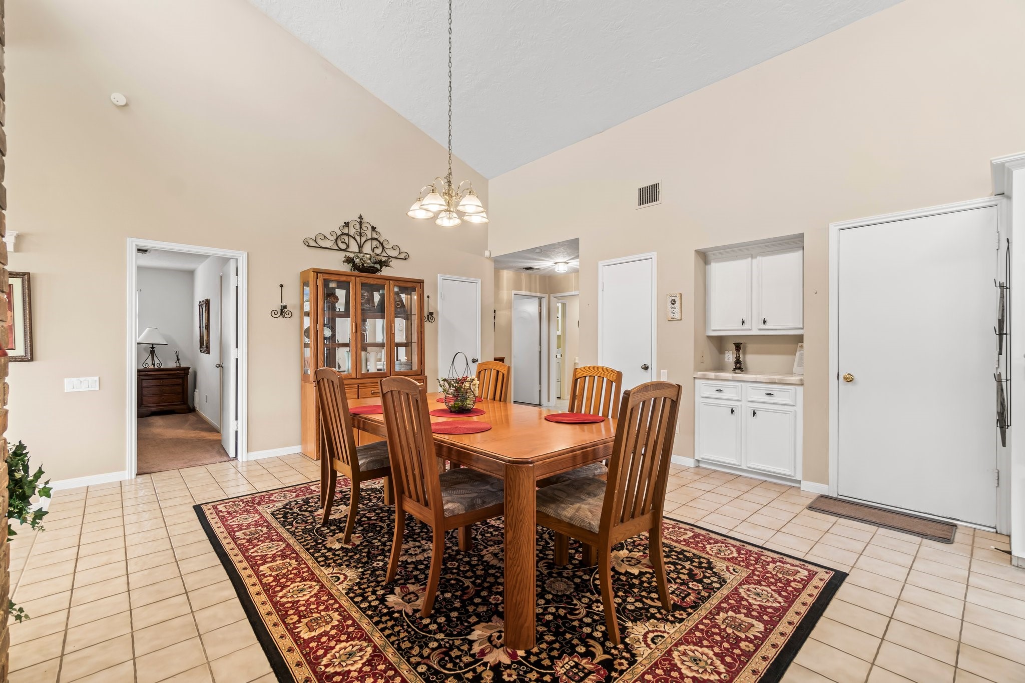 2839 Hemingway Drive Montgomery, TX 77356 - Photo 11 of 49 This photo showcases a spacious dining area with high ceilings and neutral walls. It features a wooden dining set on a decorative rug, a glass-front cabinet, and built-in storage. The space is bright and inviting, with tile flooring and an open doorway leading to another room.