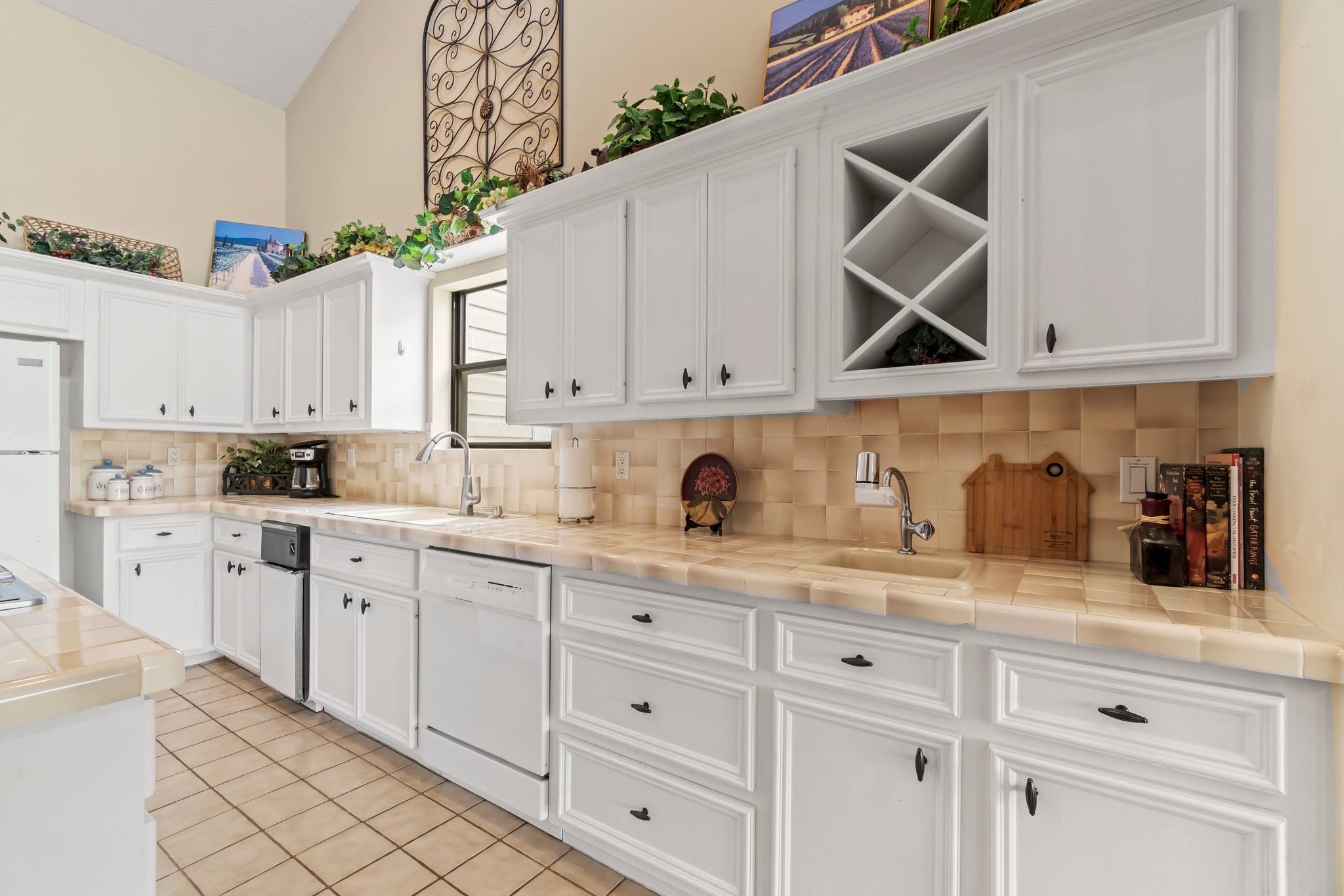 2839 Hemingway Drive Montgomery, TX 77356 - Photo 15 of 49 This is a bright kitchen featuring white cabinetry, beige tiled countertops, and a backsplash. It includes built-in wine storage, a dishwasher, and ample counter space. Decorative accents and greenery add a touch of charm.