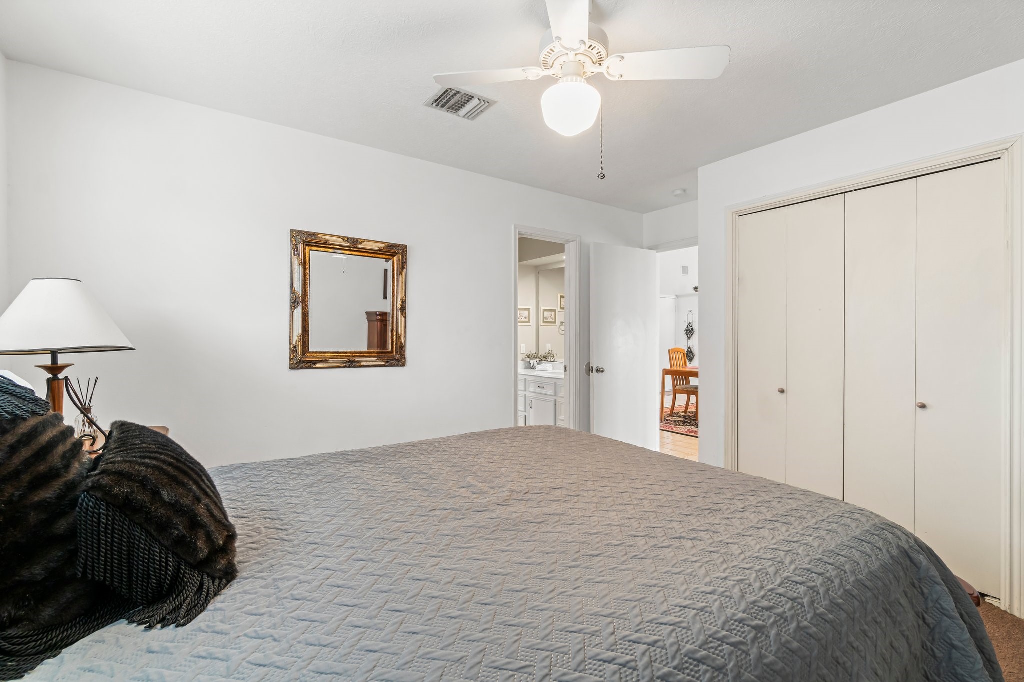 2839 Hemingway Drive Montgomery, TX 77356 - Photo 21 of 49 This photo shows a cozy bedroom with neutral walls and a ceiling fan. There's a large bed with a textured comforter, a bedside lamp, and a framed mirror on the wall. The room features a closet with double doors and an open door leading to a bathroom. Natural light brightens the space.
