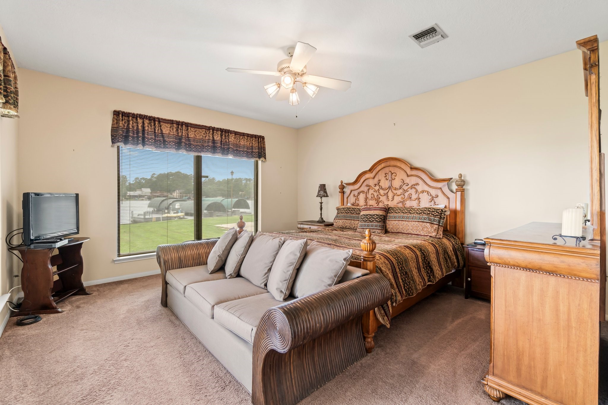 2839 Hemingway Drive Montgomery, TX 77356 - Photo 25 of 49 This spacious bedroom features a large window with natural light, a ceiling fan, and elegant furniture, including a carved wooden bed and matching dresser. The room has a cozy seating area and neutral carpet, creating a warm and inviting atmosphere.