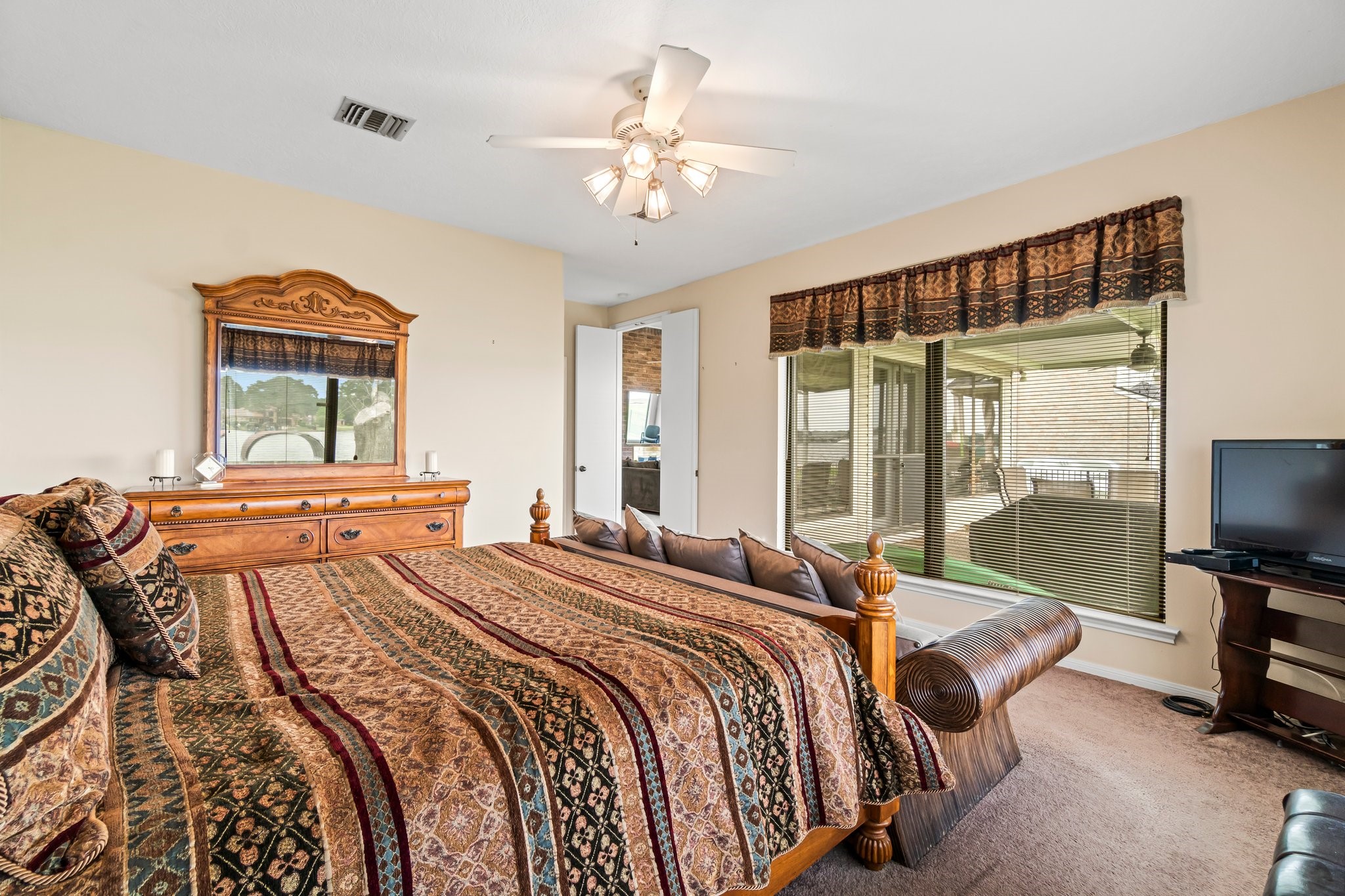 2839 Hemingway Drive Montgomery, TX 77356 - Photo 27 of 49 This cozy bedroom features a large wooden bed with patterned bedding, a matching dresser with a mirror, and a ceiling fan. There's a window with a view of a covered patio area, bringing in natural light. The room also includes a small TV on a stand.