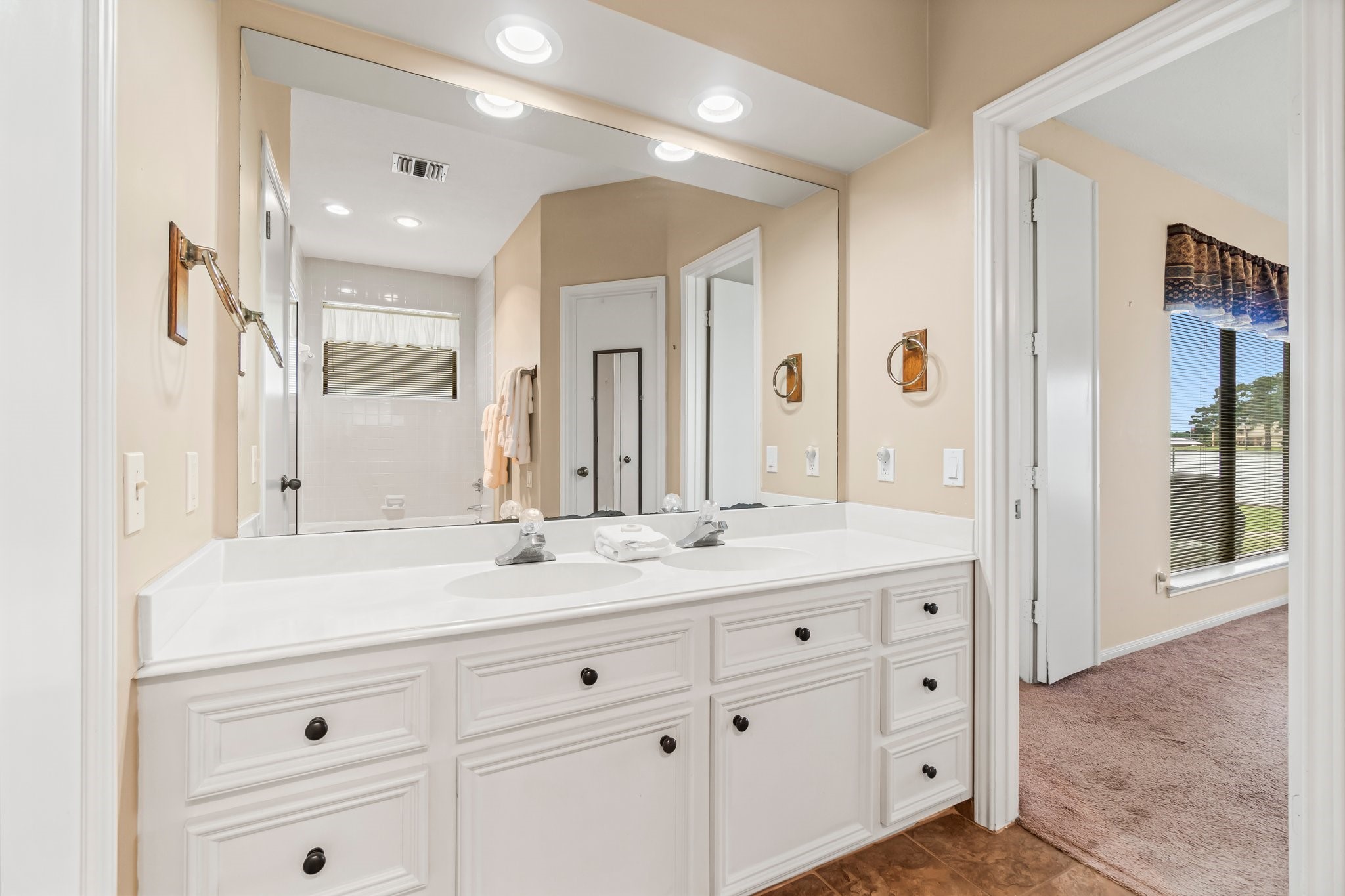 2839 Hemingway Drive Montgomery, TX 77356 - Photo 30 of 49 This photo showcases a bright bathroom with dual sinks, a large mirror, and ample cabinet space. The room has recessed lighting and leads into a carpeted area with a window, providing natural light.