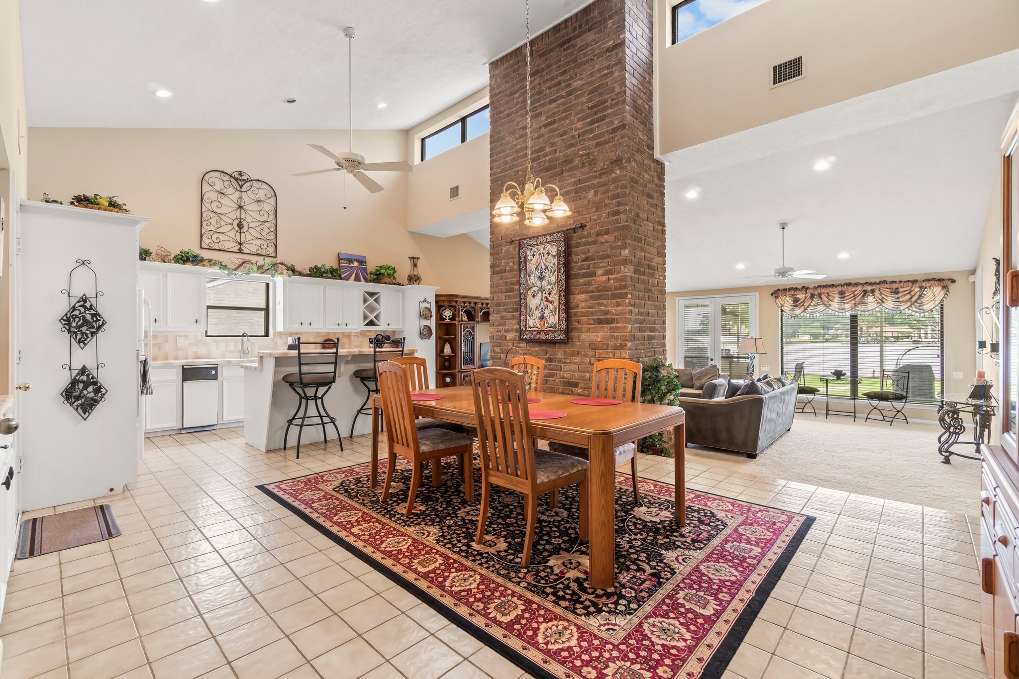 2839 Hemingway Drive Montgomery, TX 77356 - Photo 9 of 49 This spacious open-concept living area features a high ceiling with abundant natural light, a central brick column, and a seamless flow from the kitchen to the dining and living spaces. The kitchen offers white cabinetry and a breakfast bar, while the living room provides a cozy, inviting atmosphere with large windows overlooking the outdoors.
