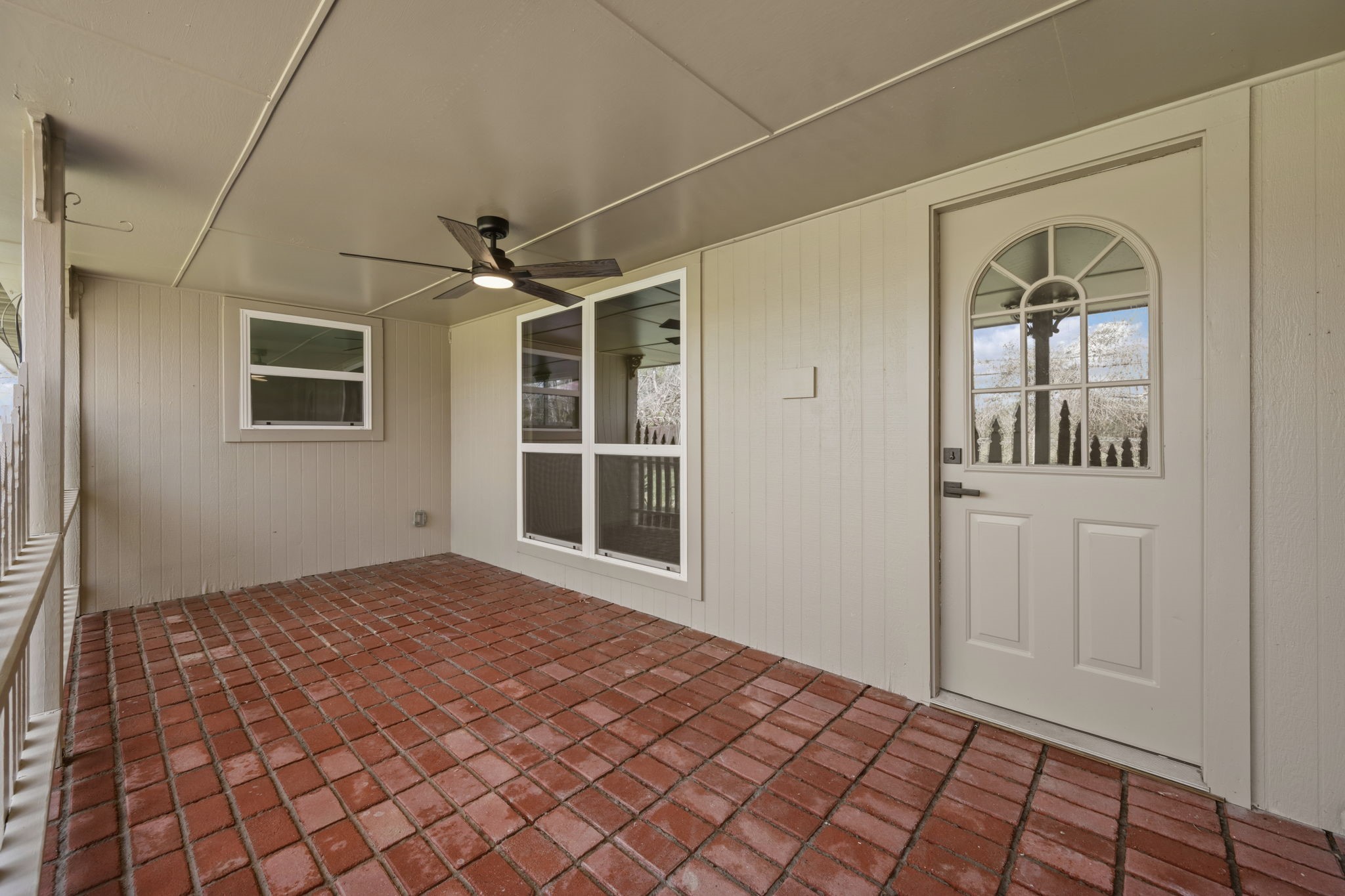 210 Ridgemont Drive Alvin, TX 77511 - Photo 24 of 27 This photo shows a covered patio with red brick flooring, light-colored paneled walls, and a ceiling fan. There are two windows and a door with a decorative window, creating a cozy and functional outdoor space.