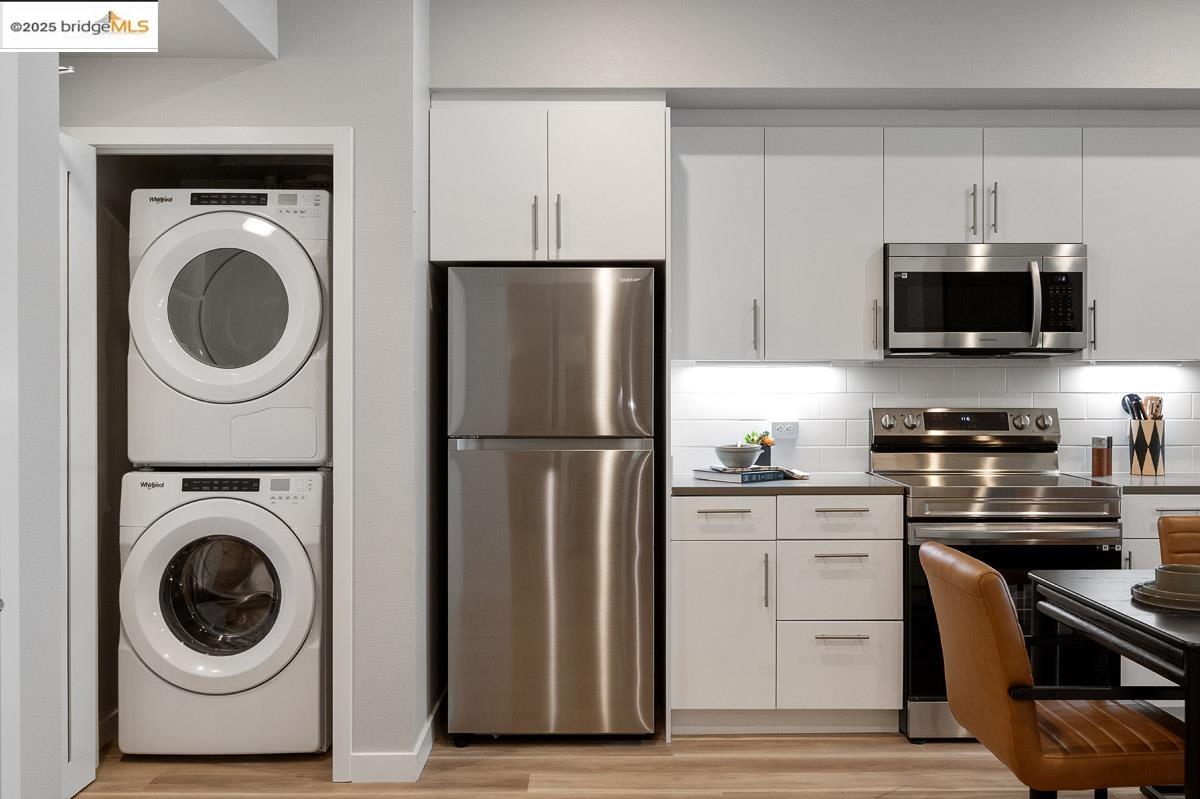 260 Brooklyn Basin Way, Unit 428 Oakland, CA 94606 - Photo 14 of 44 Kitchen featuring stainless steel appliances, white cabinetry, stacked washer and clothes dryer, backsplash, and light wood-style floors