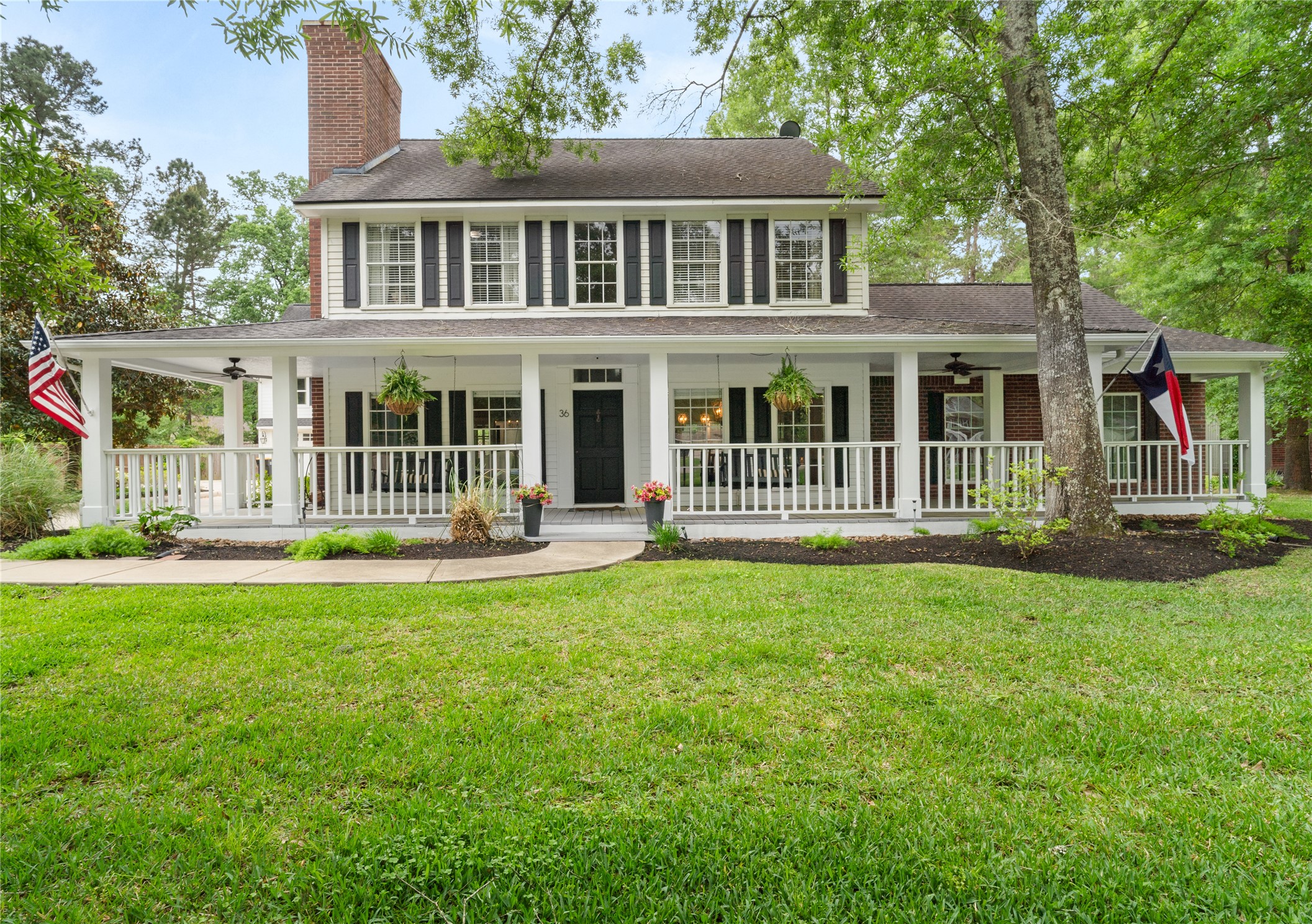 36 Wind River Drive Conroe, TX 77384 - Photo 1 of 45 This charming two-story home features a classic white exterior with black shutters, a spacious wrap-around porch, and a lush, well-maintained front yard. The porch is adorned with hanging plants and flags, offering a welcoming and picturesque setting.