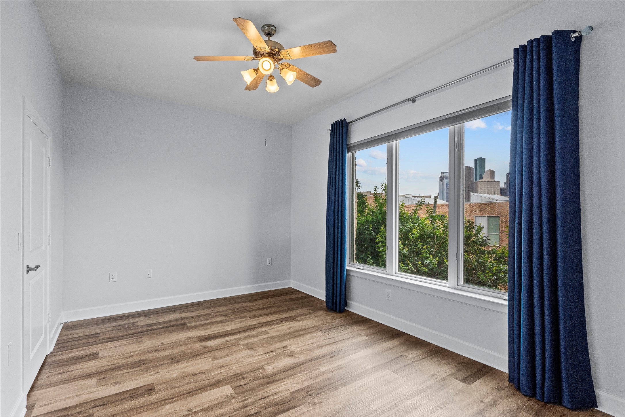 1900 Genesee Street, Unit 203 Houston, TX 77006 - Photo 17 of 30 This bright room features large windows with blue curtains, offering plenty of natural light and a view of the cityscape. The space has sleek wood flooring and a ceiling fan, creating a modern and comfortable atmosphere.