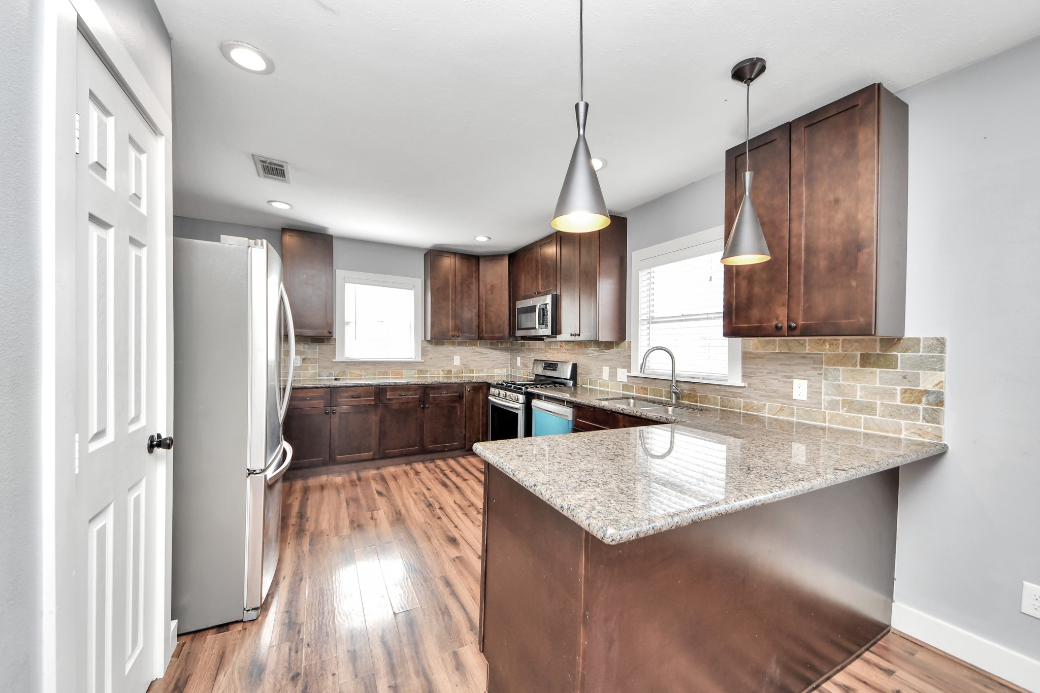 2820 Holman Street Houston, TX 77004 - Photo 15 of 38 Modern kitchen with granite countertops, stainless steel appliances, and dark wood cabinetry. Features a spacious layout with a breakfast bar and pendant lighting, complemented by wood flooring and a stylish tile backsplash.