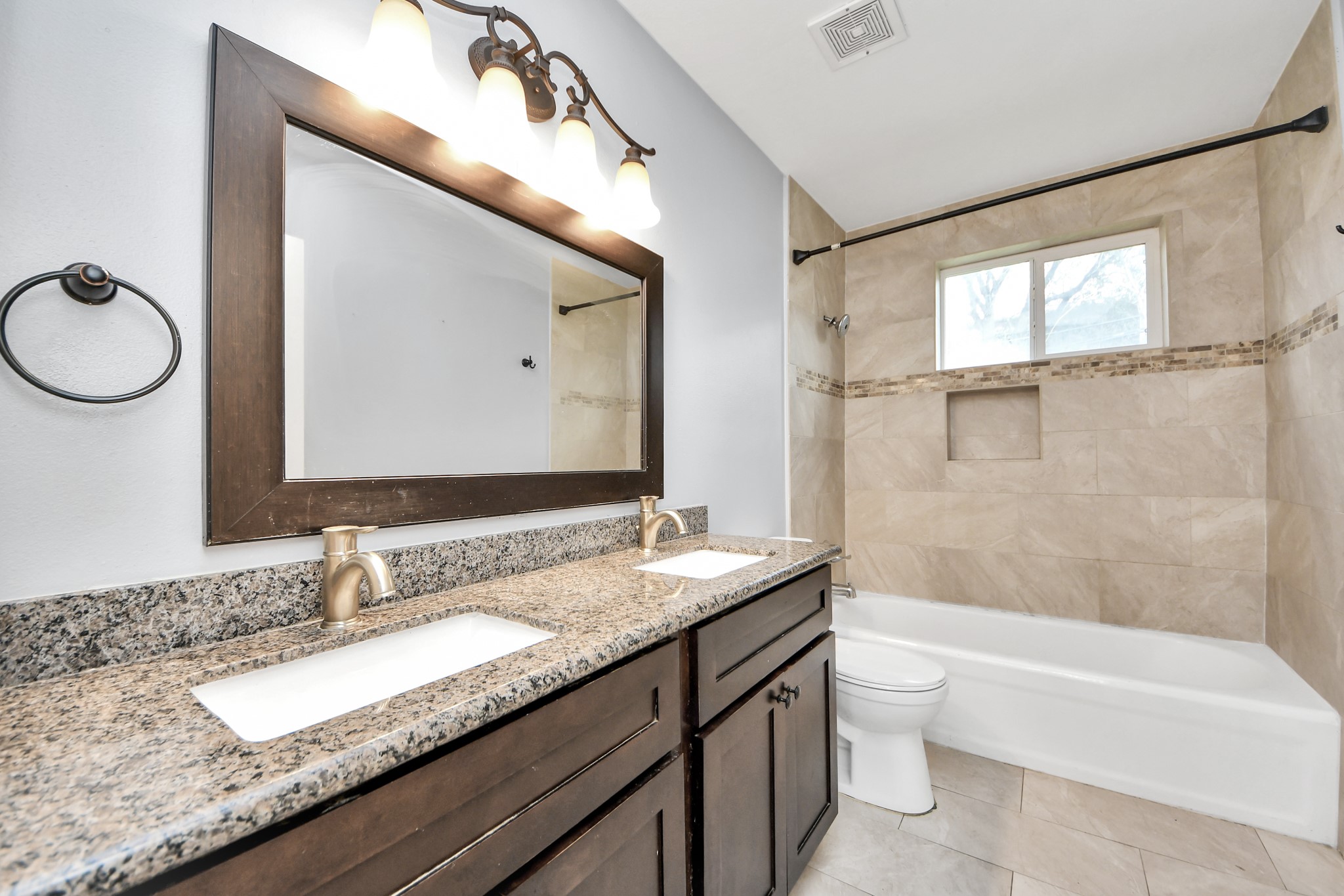 2820 Holman Street Houston, TX 77004 - Photo 24 of 38 This bathroom features a dual-sink vanity with granite countertops, elegant lighting, and a large framed mirror. The bathtub is set against a tiled wall with a decorative border and includes a window for natural light, creating a bright and inviting space.
