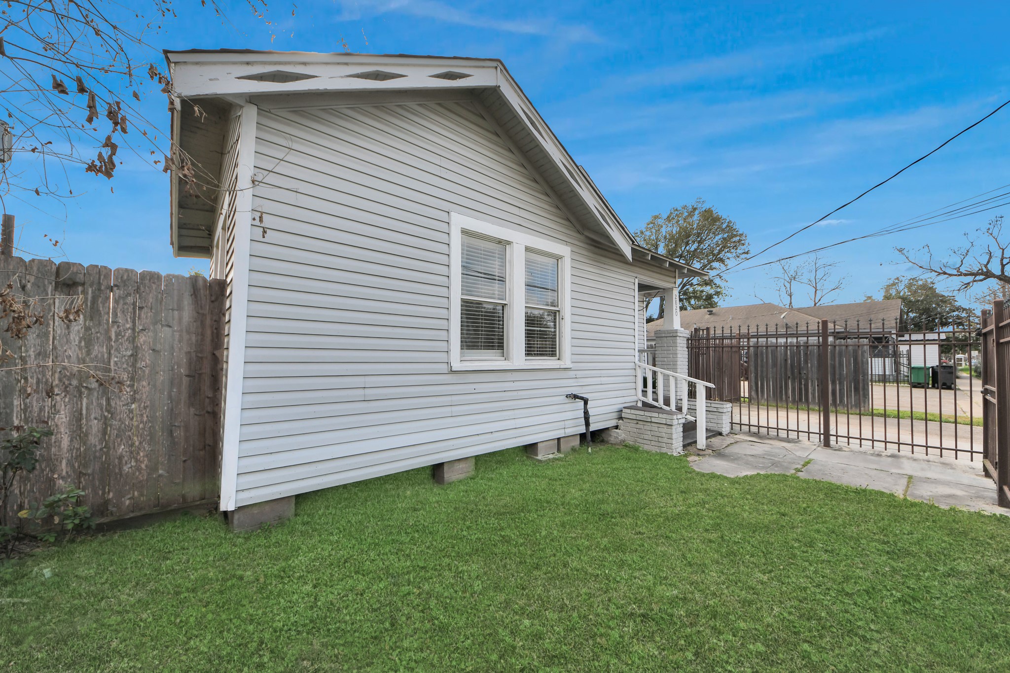 2820 Holman Street Houston, TX 77004 - Photo 30 of 38 This photo shows the side of a quaint house with gray siding, featuring a small porch and a fenced yard. The property is bordered by a wooden fence and a gated driveway, providing a sense of privacy.