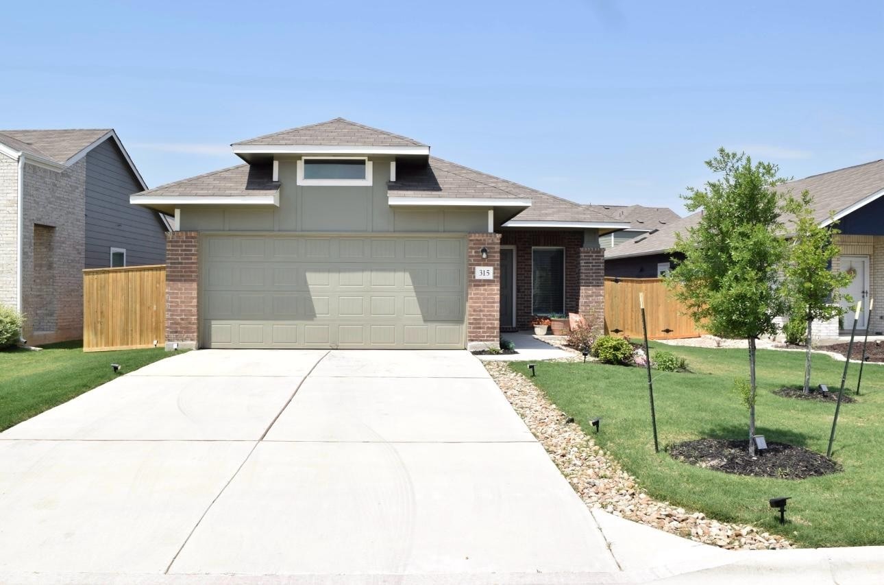 View of front facade with a garage, a shingled roof, driveway, brick siding, and fence